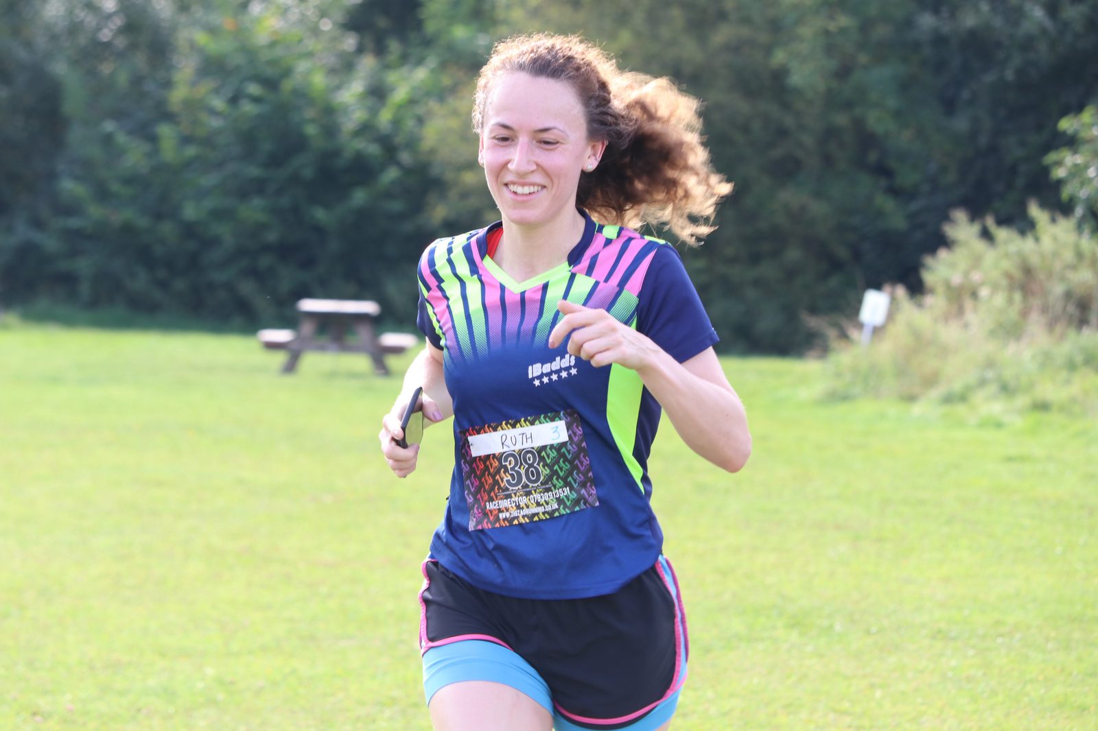 A woman with curly hair wearing a colorful athletic shirt and running shorts is running on a grassy area. She is smiling and holding what appears to be a small object. The background shows greenery and a blurred picnic table.
