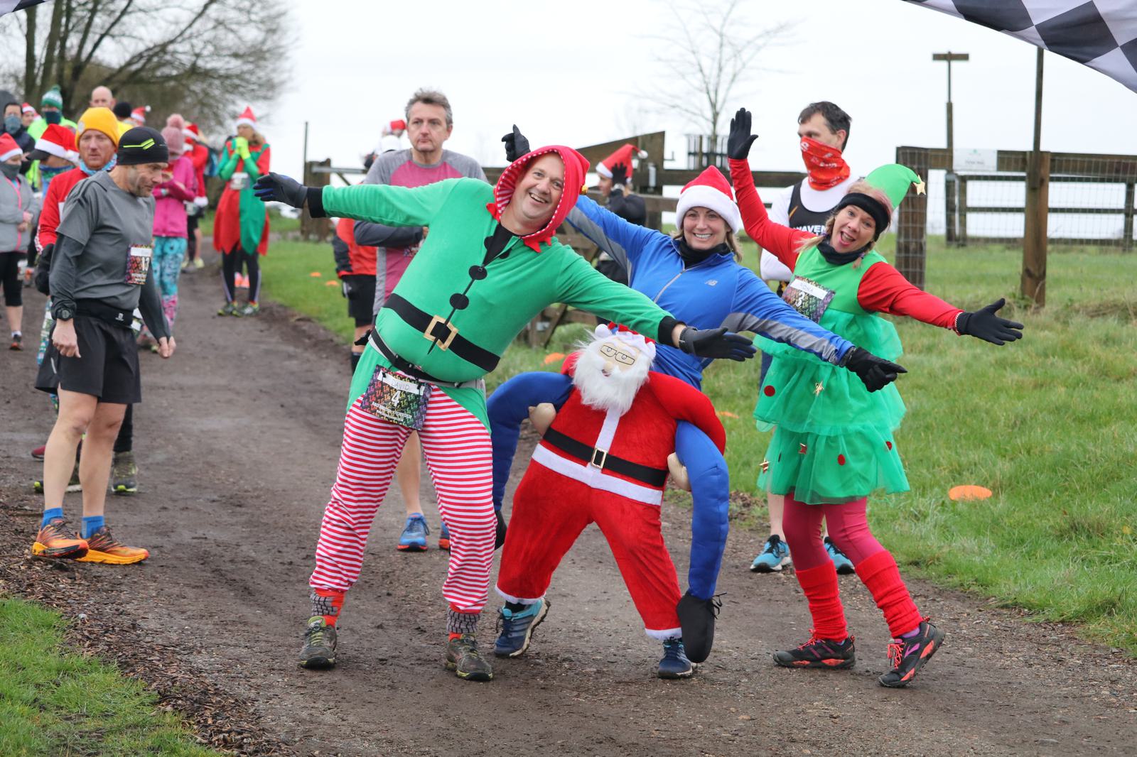 A group of runners in festive costumes, including an elf, Christmas tree, and Santa, pose cheerfully for the camera on a rural trail during a race. Other runners in holiday attire are visible in the background. The mood is joyful and energetic.