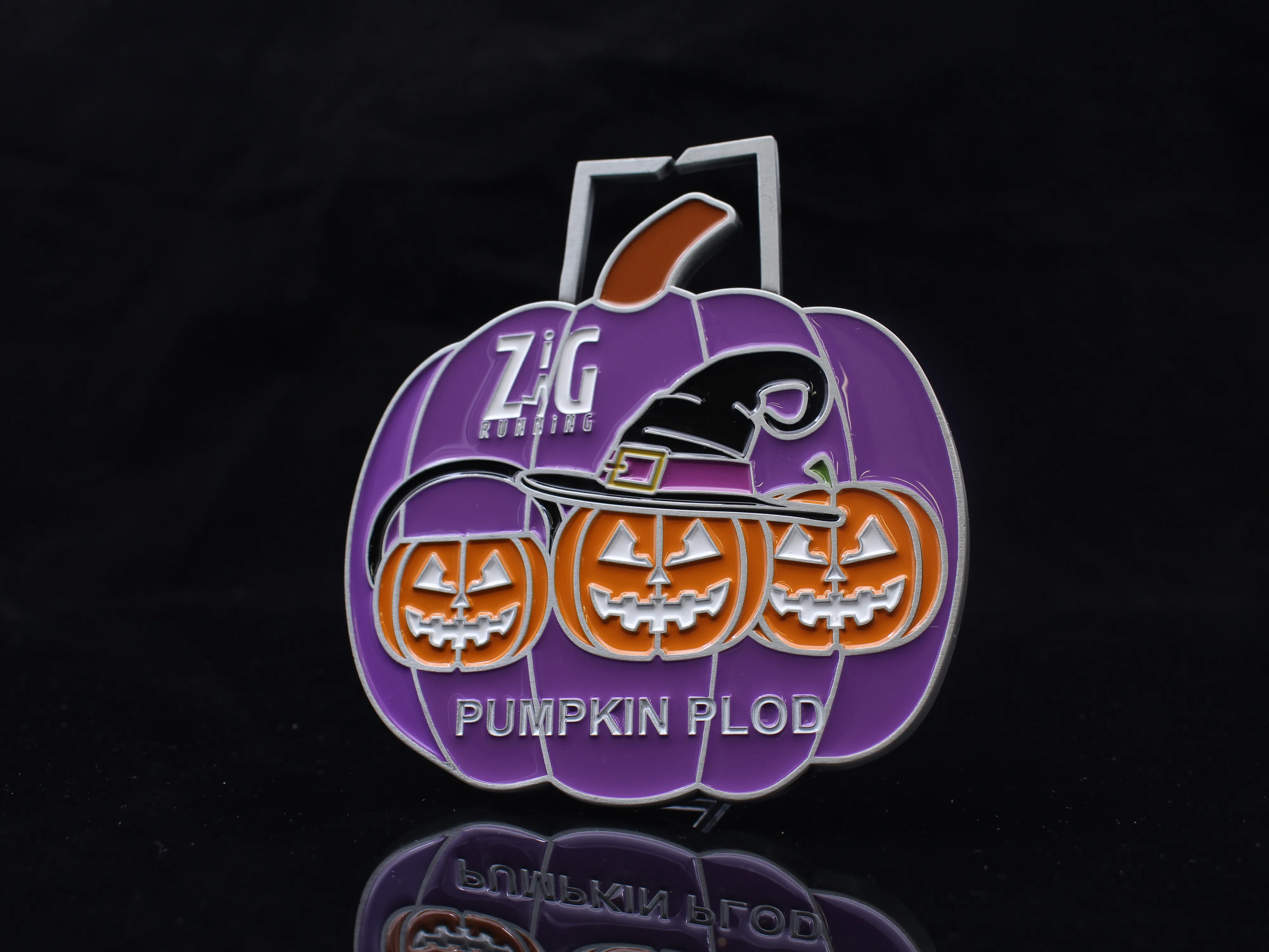 A Halloween-themed medal shaped like a purple pumpkin with three orange jack-o’-lantern faces, one wearing a witch hat, and text reading “Zig Running Pumpkin Plod” on a black background.