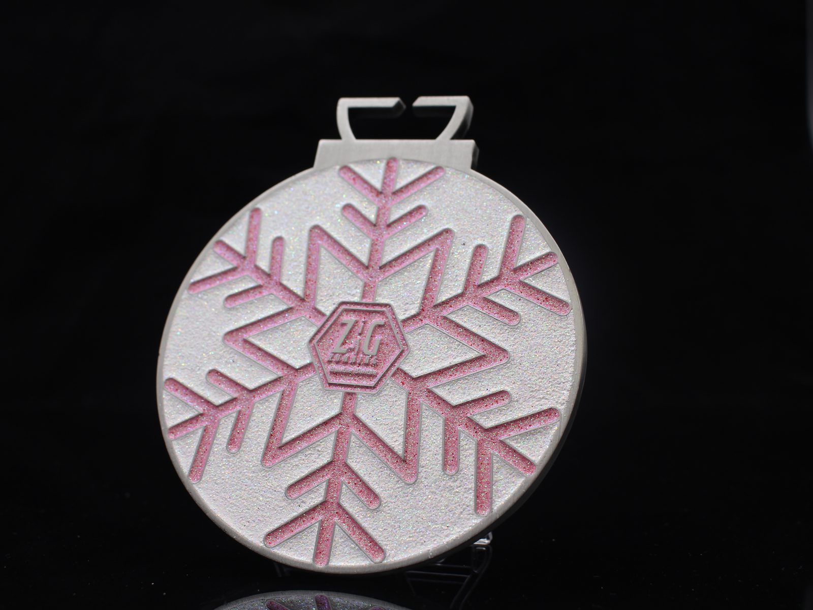 A round, glittery white ornament with a pink snowflake design and a small hexagon logo in the center, set against a black background.