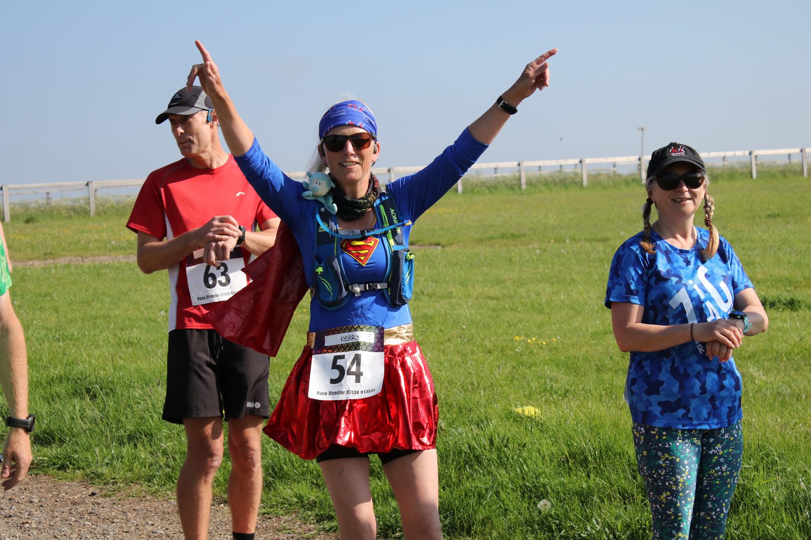 A runner dressed in a blue and red superhero costume, with race bib number 54, raises her arms in celebration. She is standing between two other participants, a man in a red shirt with bib number 63 and a woman in a blue camouflage shirt with bib number 110. They are outdoors on a sunny day with a grassy field in the background.