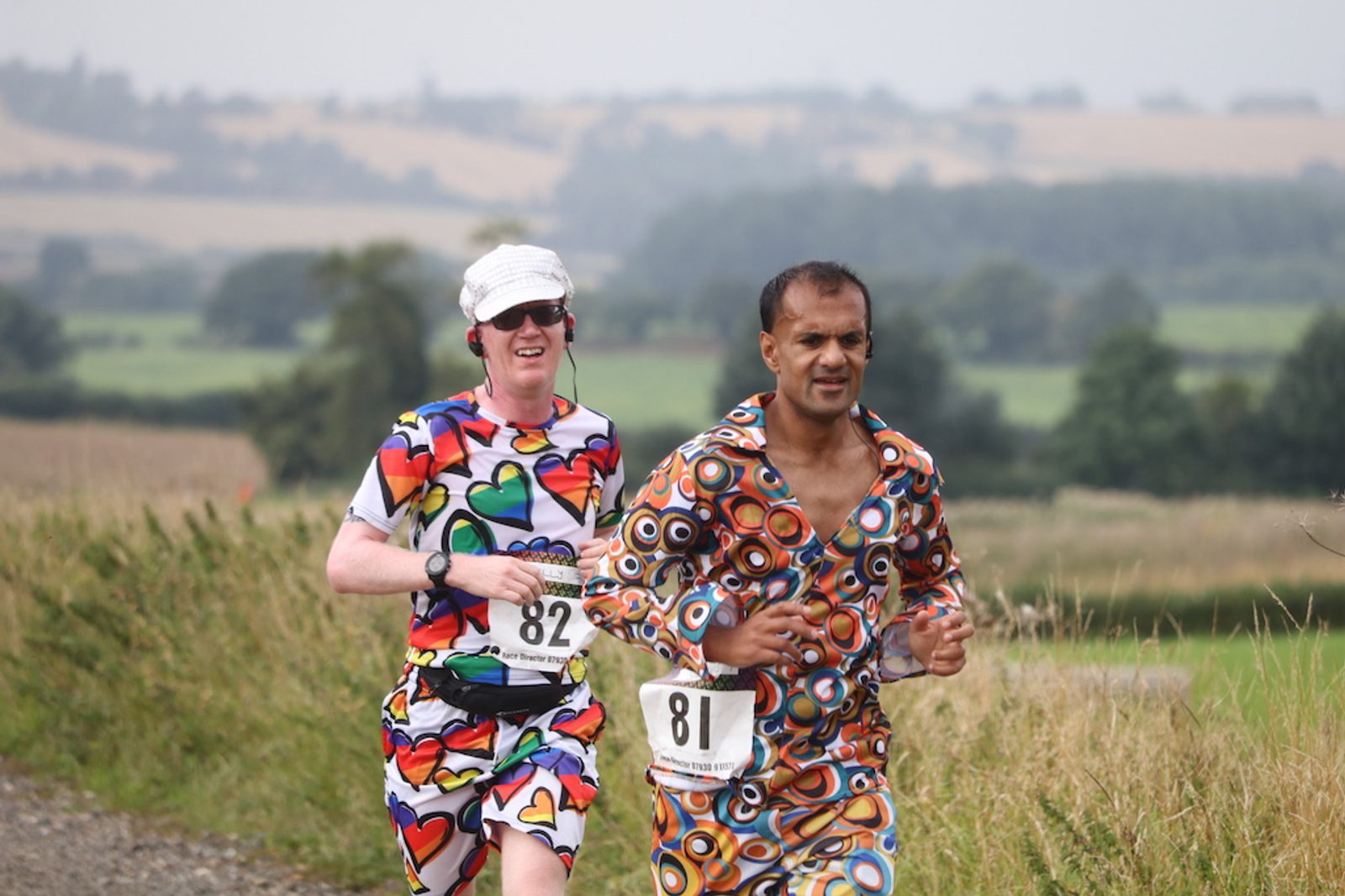 Two runners dressed in colorful, patterned outfits compete in a race on a rural path. Runner 81, in a brown, orange, and blue geometric outfit, is ahead of runner 82, who wears a white cap and a top with multicolored heart designs. Countryside scenery is visible in the background.