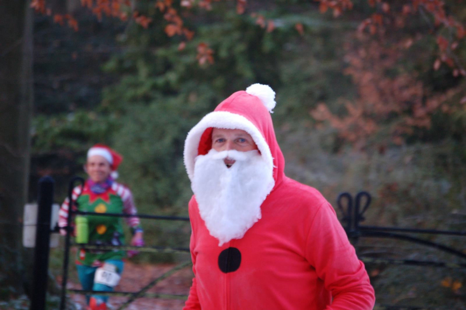 A person dressed as Santa Claus, complete with a red suit and white beard, jogs outside. Another person in the background is dressed in a colorful elf costume with a pointy hat, green outfit, and Christmas-themed attire. Trees with autumn leaves surround the scene.