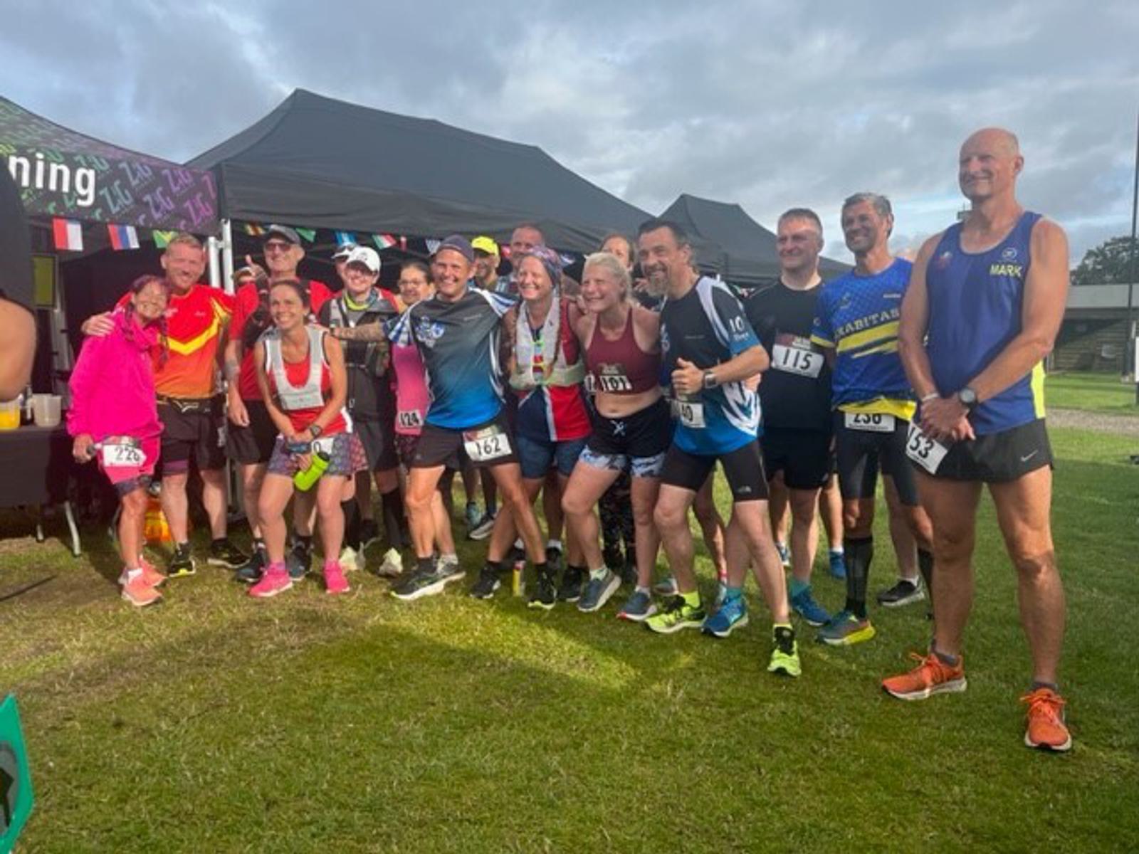 A group of runners, wearing race numbers, stands together and smiles for a photo under a tent on a grassy field. Some runners are holding medals. Everyone appears happy and energetic, suggesting they have just finished a race or event.