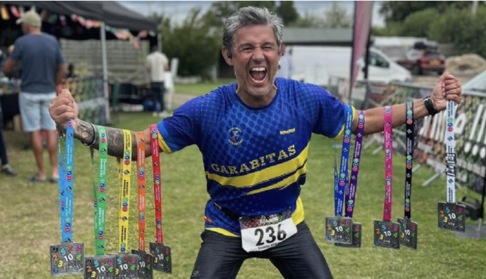 A man is smiling broadly and flexing his arms while holding a series of colorful medals in each hand. He is wearing a blue and yellow sports jersey with the word "GARABITAS" and a race number "236" on his waist. There are people and tents in the background.