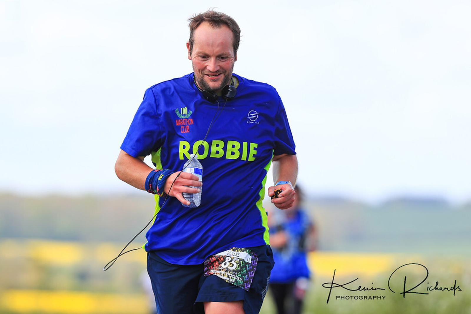 A runner wearing a blue shirt with the name "ROBBIE" printed on it, holding a water bottle, and smiling during an outdoor race. The landscape in the background is slightly blurred, showing green fields and a cloudy sky. The logo "Kevin Richards Photography" is visible in the corner.