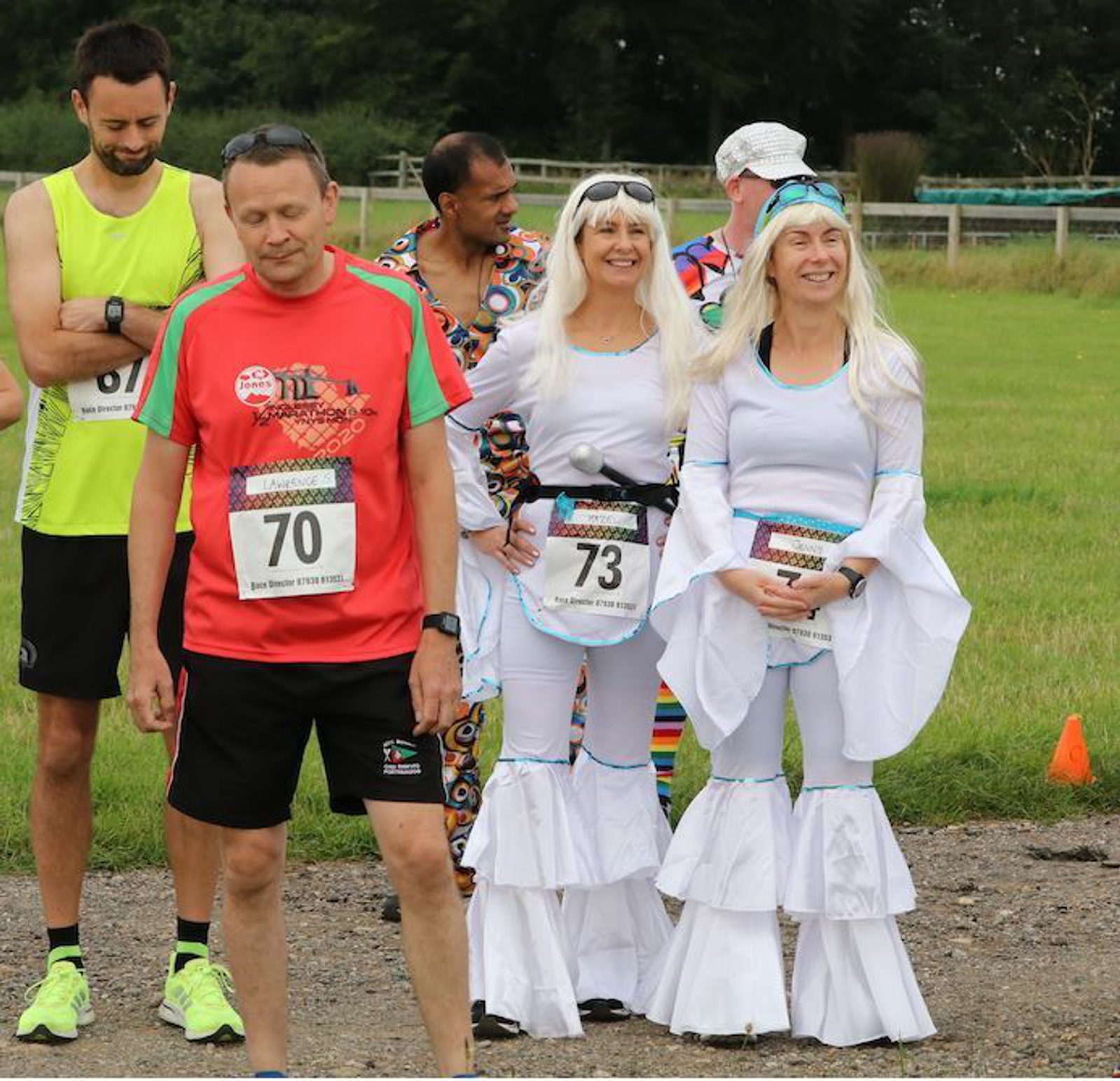 A group of people preparing for a race. Three women dressed in white costumes with long blonde wigs and numbered bibs, 73 and 70, stand smiling. Two men in running gear and numbered bibs are nearby, one in a green vest with bib 87 and the other in a red shirt.