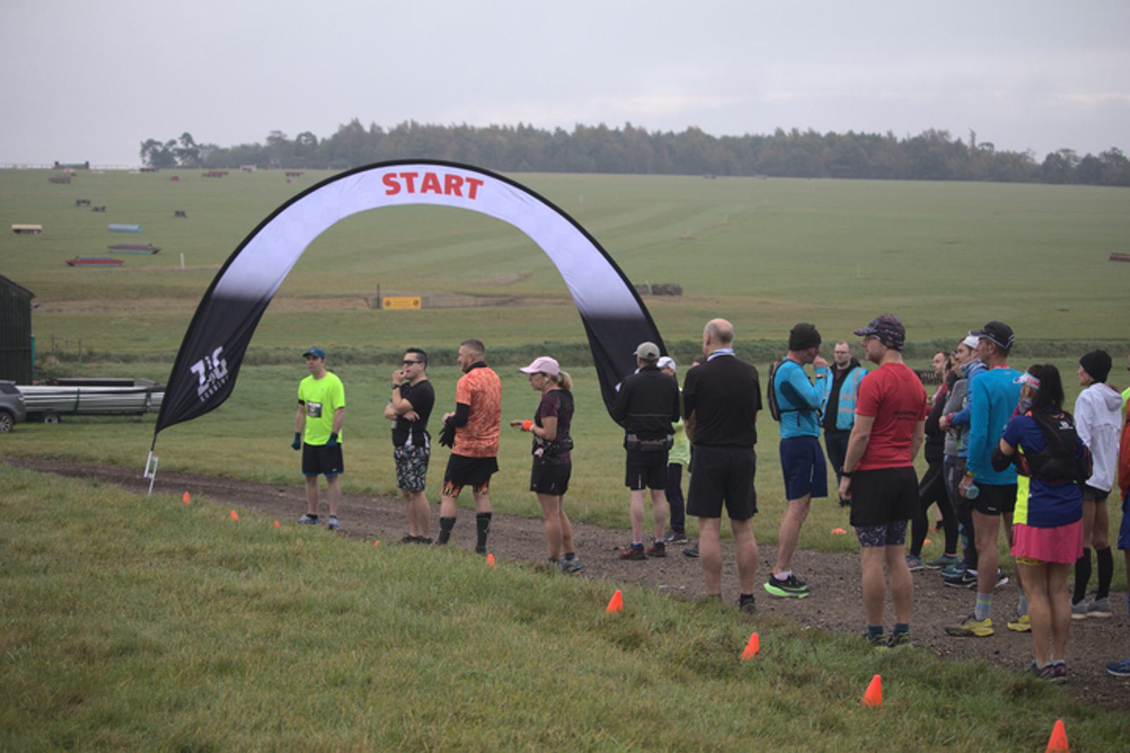 A group of runners gather near a "START" banner at the beginning of an outdoor race on a grassy field. Some are chatting while others are warming up. Orange cones mark the start area, and a vast open field stretches out in the background under a cloudy sky.