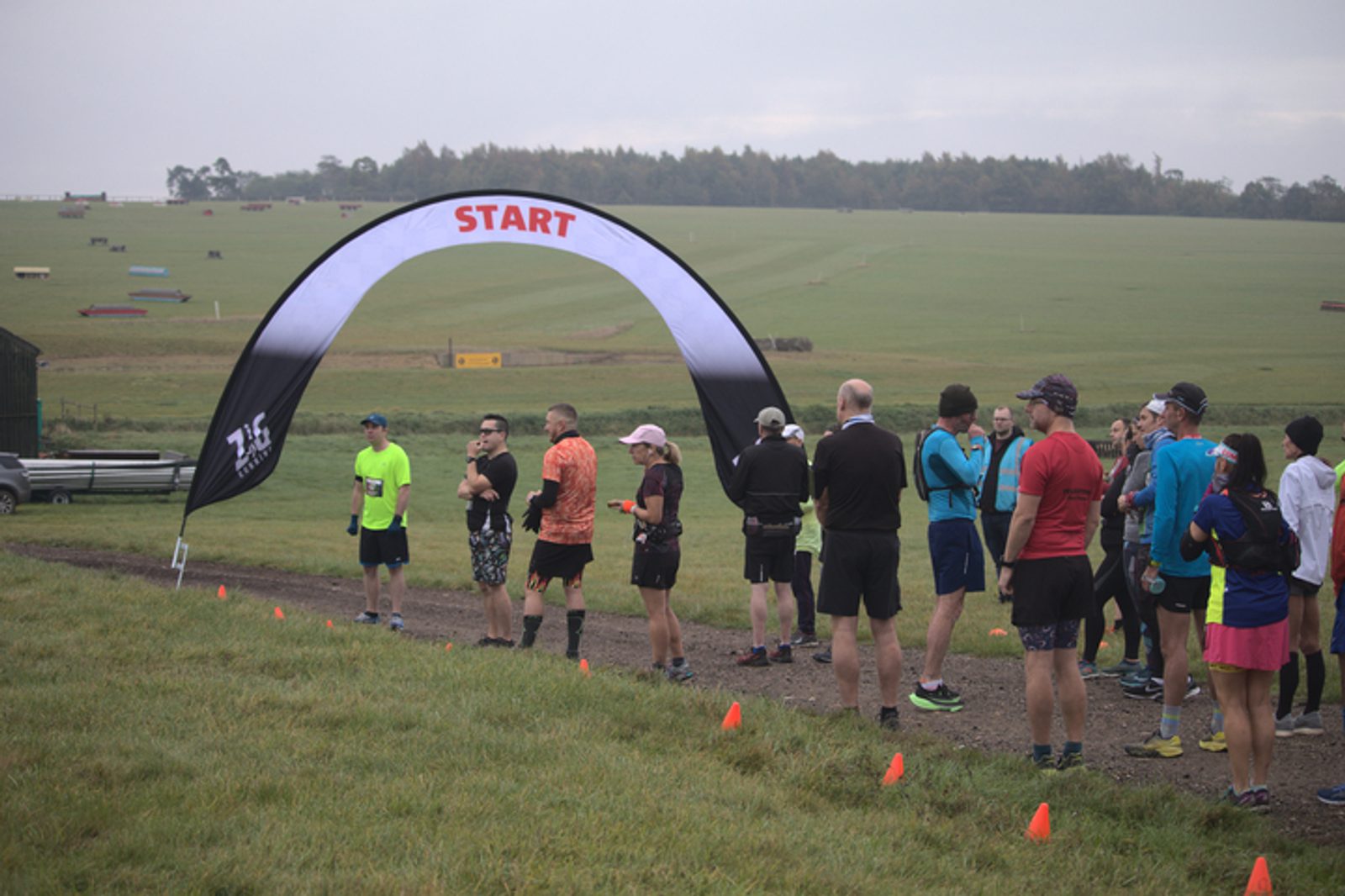 A group of runners gathers under a black and white arch marked "START" at the beginning of a race. The runners are dressed in various athletic attire and stand on a gravel path with grassy fields and a wooded area in the background. Orange cones line the path.