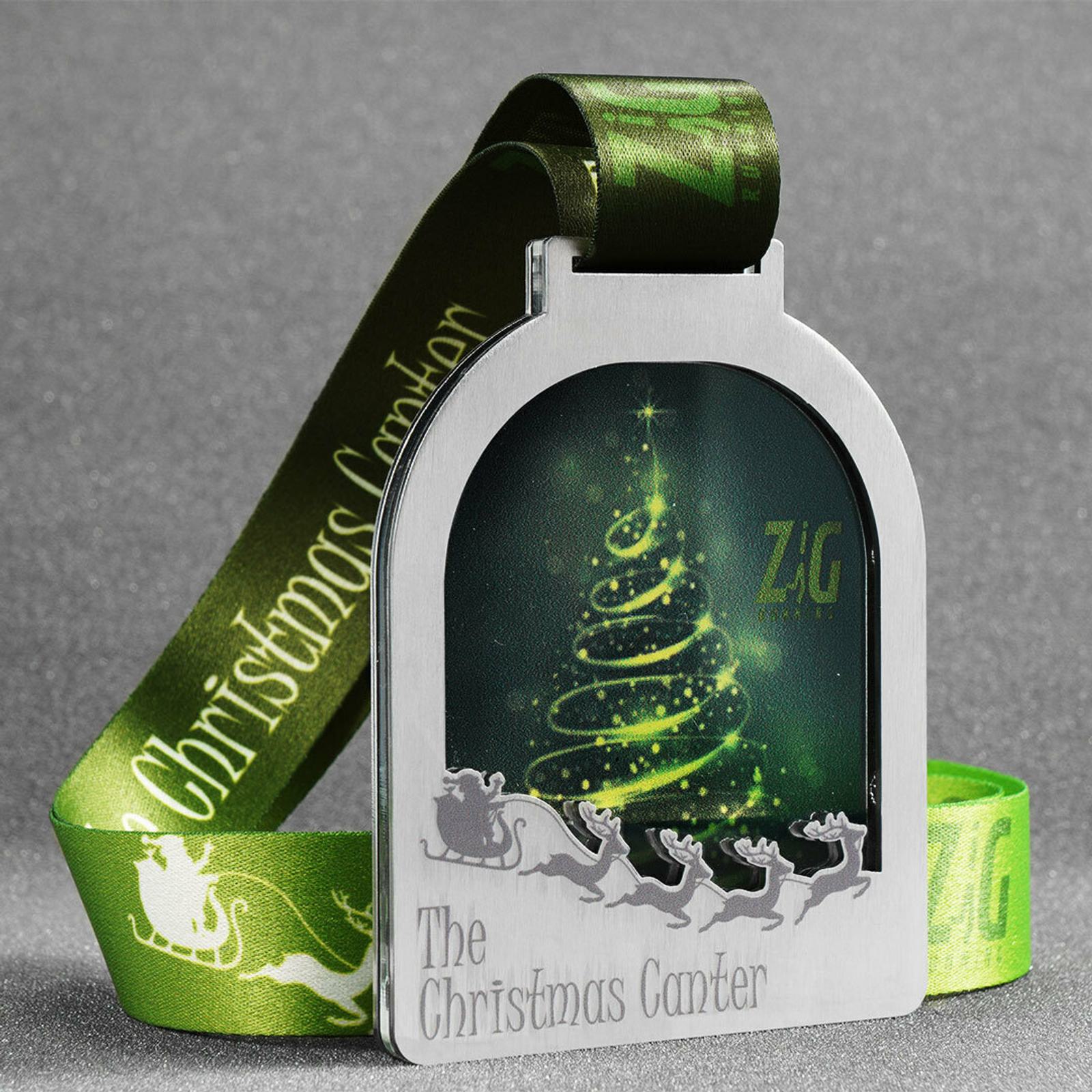 A silver medal with a green ribbon featuring text "Christmas Canter." The medal has a cutout window showing an illuminated stylized Christmas tree and the letters "ZIG." Below the window, silhouettes of Santa's sleigh and reindeer are visible.