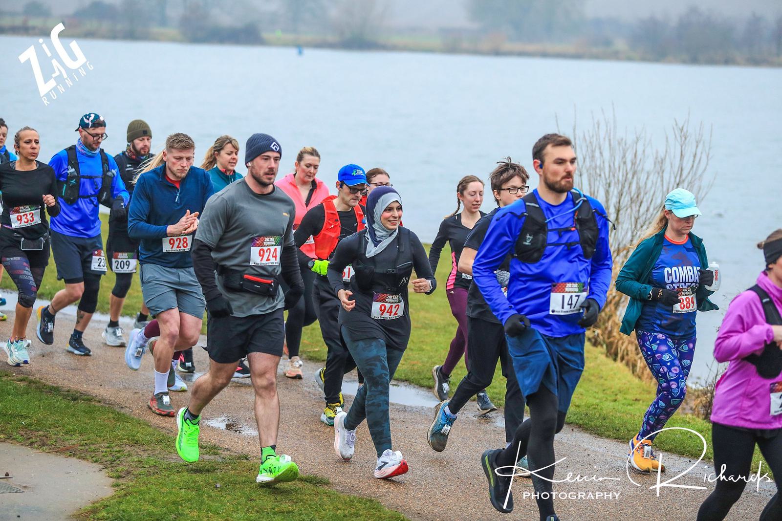 A group of runners in athletic gear jogging along a lakeside path during a race. Some wear race bibs with numbers, and a few are dressed in bright colors. The sky is overcast, and there is water and vegetation in the background.