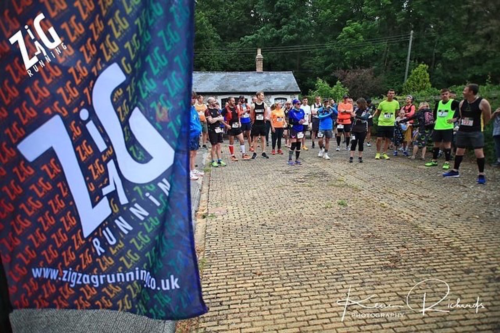 A group of runners stand in a semicircle, listening to instructions before a race. In the foreground, a banner reads "Zigzag Running" along with their website. Around them, there are trees and a small building. The ground is cobbled. Photography credit is visible.