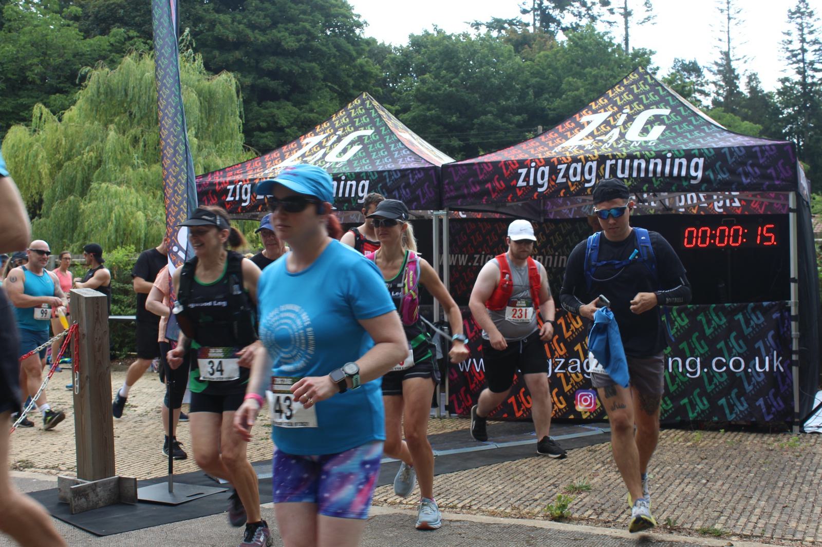 A group of runners is starting a race outside, passing under tents labeled "zig zag running." Each runner wears a numbered bib. A digital timer shows 00:00:15. Trees and a weeping willow are in the background.