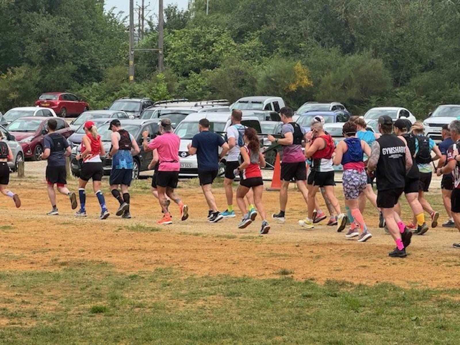A group of people in athletic clothing are running together on a grassy field near a parking lot filled with cars, with trees in the background.