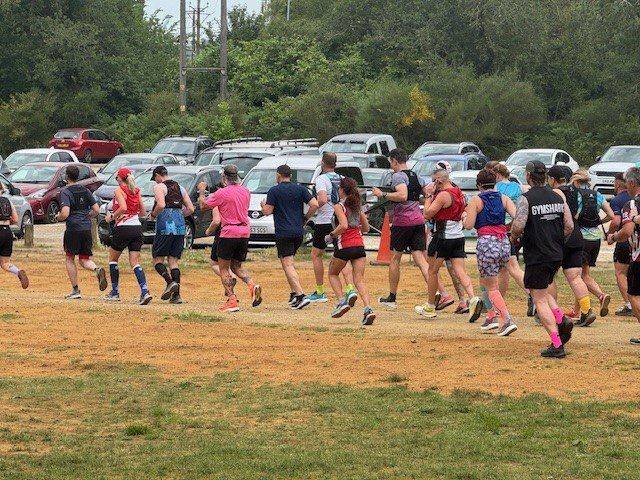 A group of people in athletic clothing are running together on a grassy field near a parking lot filled with cars, with trees in the background.