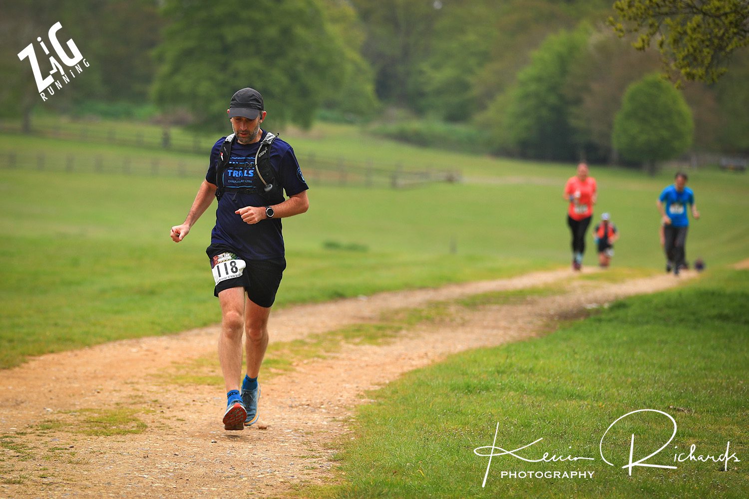 A man in a black cap and blue shirt runs on a dirt path with a number bib (118) pinned to his shorts. Behind him, three runners move in the same direction. In the background, there are trees and green grass. The image has a watermark for "Kevin Richards Photography" and "Zig Running.