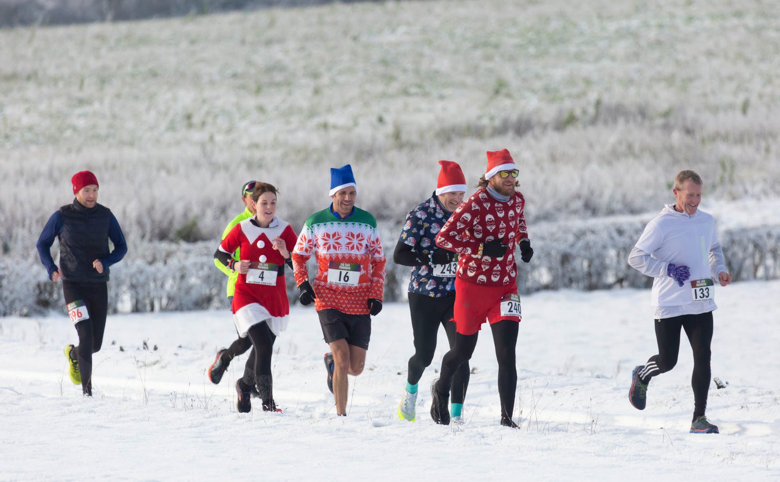 A group of runners in festive attire, including Santa hats and holiday sweaters, participate in a winter race on a snowy path. Bib numbers are visible on their clothing, and the background shows a frosty landscape with fields and sparse bushes.