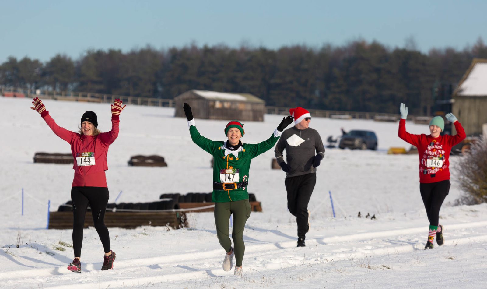 Four runners are making their way along a snowy path. The runner in the center wears a festive green costume and is smiling with raised arms. The others are dressed warmly, two in Santa hats, and all appear to be enjoying the run, with a snowy landscape in the background.