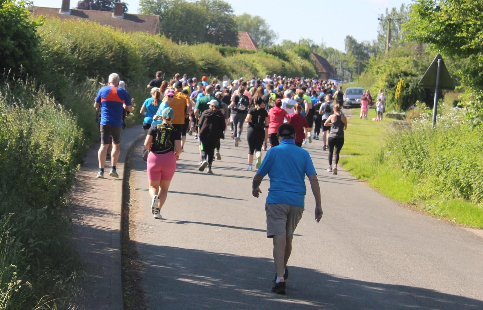 A large group of people participate in a running event on a sunny day, moving along a rural road lined with green bushes and trees. Many are dressed in colorful athletic wear, and the road sign indicates a gentle curve ahead.