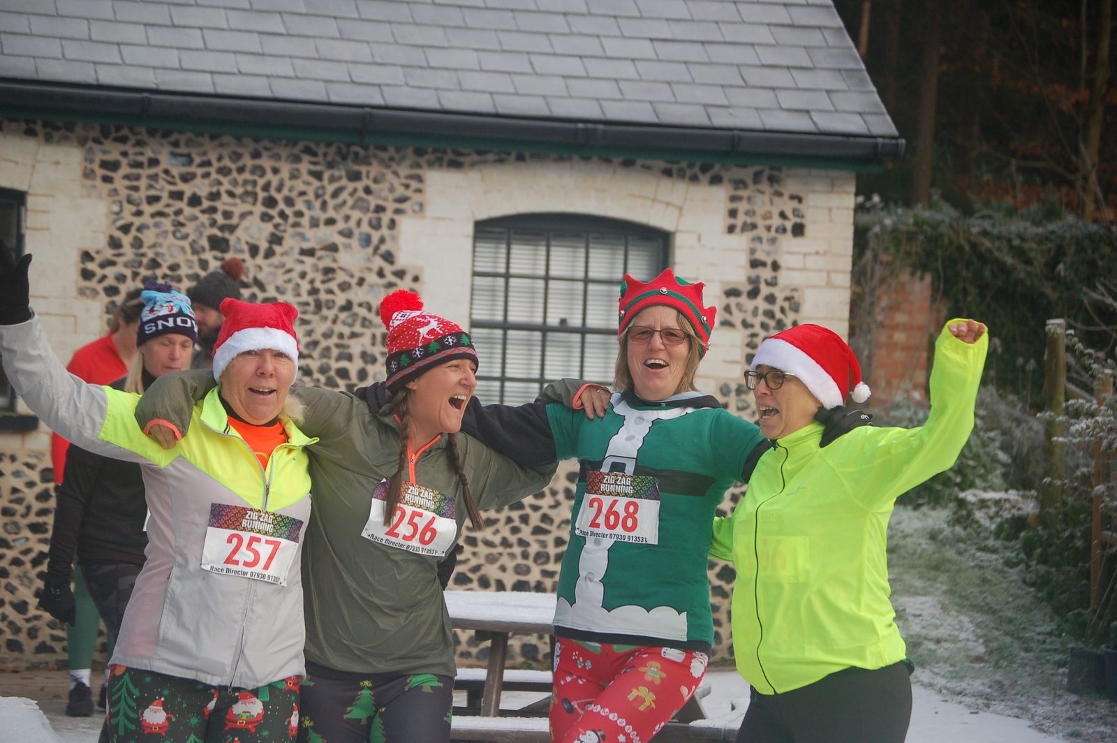 Four people dressed in festive running attire, including Santa hats and an elf costume, celebrate with arms around each other in front of a building with brick and stone walls. They wear race bibs numbered 256, 257, and 268, suggesting participation in a holiday-themed race.