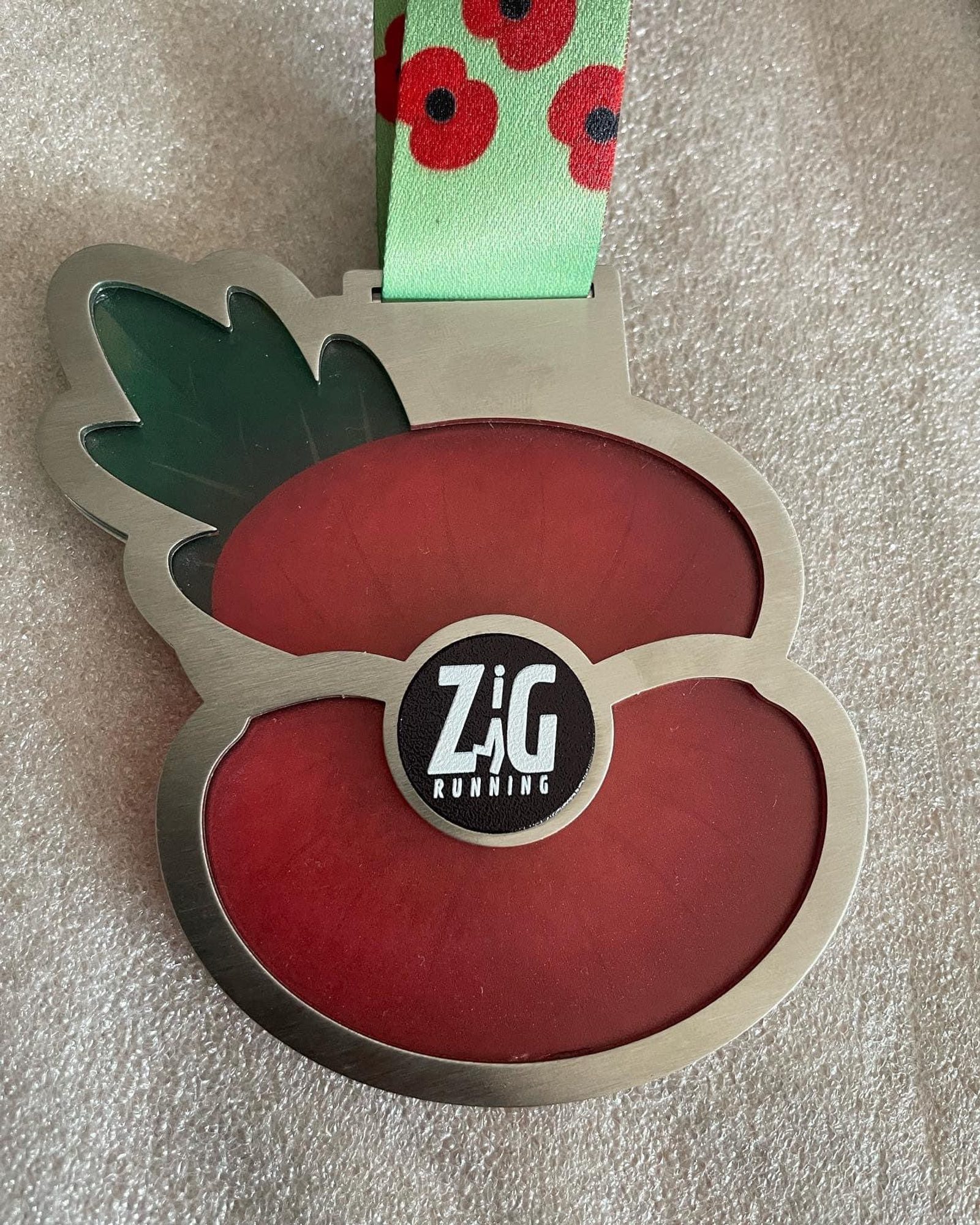 A close-up view of a medal shaped like a poppy flower, featuring a green leaf. The medal has "Zig Running" written in black and white at the center. The attached ribbon is light green with a pattern of red poppies. The background is a light-colored fabric.