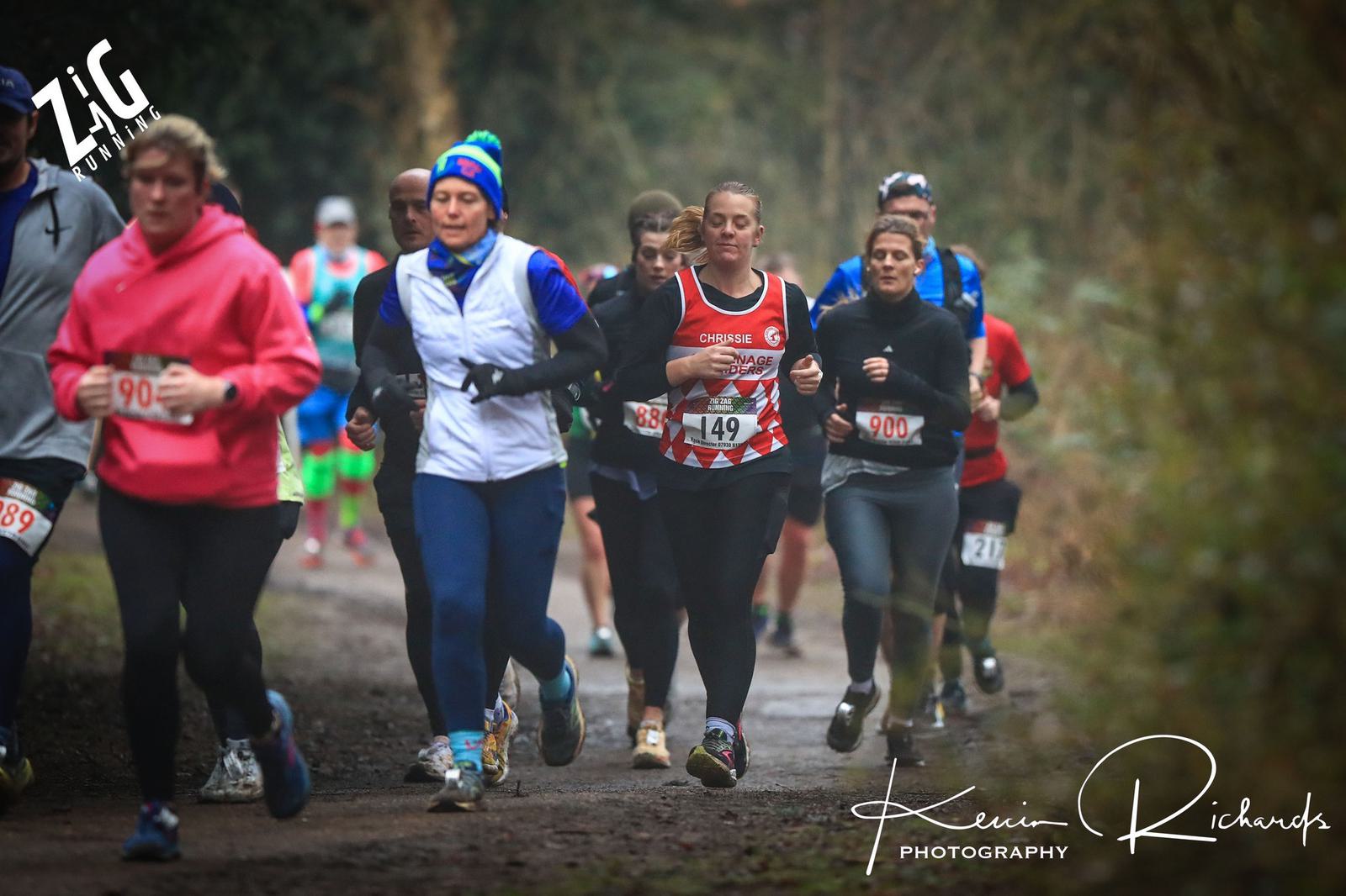 A group of runners participates in an outdoor race, moving along a forest path. They wear various athletic attire, with visible race numbers. The environment is misty and the path is surrounded by trees.