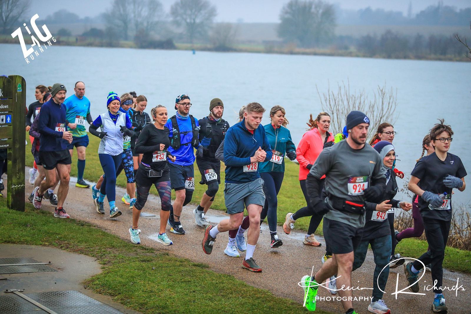 A group of runners in athletic gear participate in an outdoor race on a path beside a lake. The sky is overcast, and leafless trees are in the background. Some runners wear numbered bibs and hats.