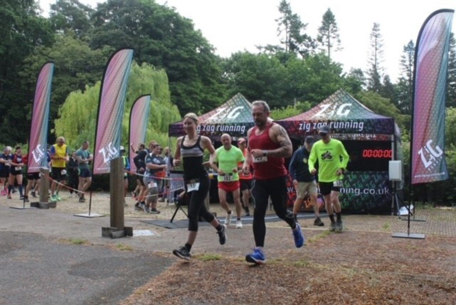 A group of runners starting a race, with some wearing race bibs. They are running past colorful flags and a Zig Zag Running event tent surrounded by trees. A small digital timer is in the background. The atmosphere appears lively and energetic.