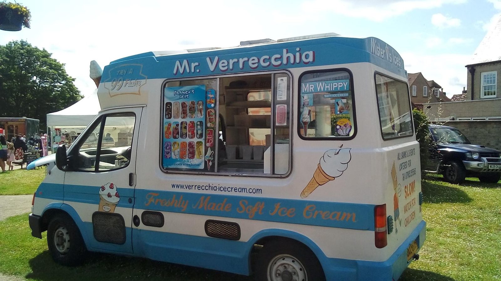 A blue and white ice cream van labeled "Mr. Verrecchia" parked on grass. The side displays various ice cream options and prices. The front menu board reads "Freshly Made Soft Ice Cream." A small ice cream cone illustration and website are visible on the van.