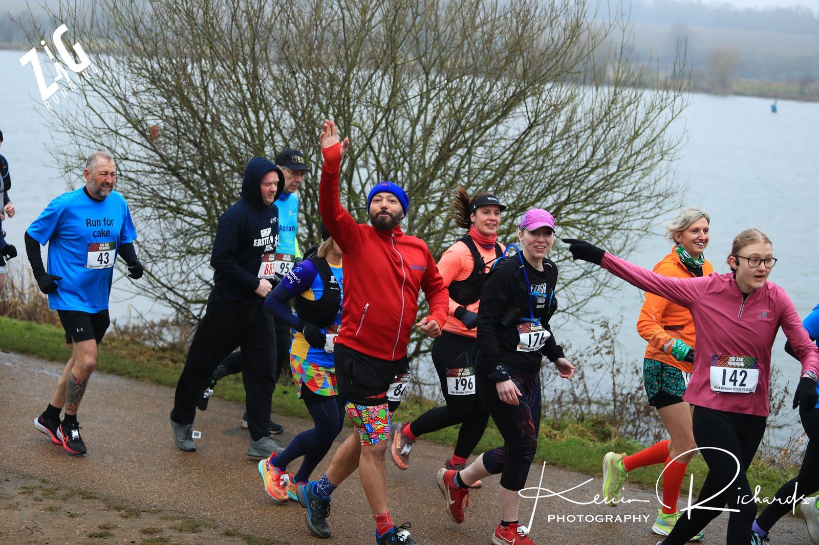 A group of diverse runners, bundled in vibrant winter gear, jogs enthusiastically along a lakeside trail. A man in a red jacket raises his arm cheerfully. Bib numbers visible, including 142. Overcast sky and bare trees set a chilly atmosphere.
