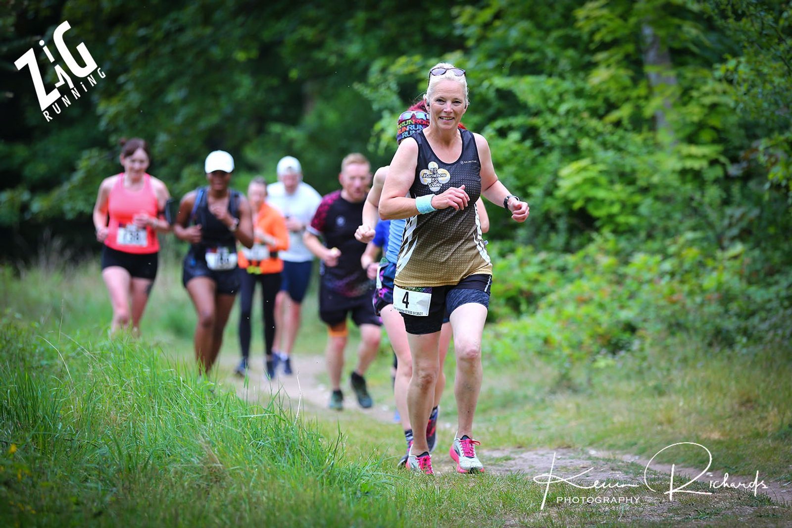 Group of runners on a forest trail, led by a woman wearing a tank top and shorts with the number 4 bib. Other runners follow closely behind her. The trail is surrounded by lush green foliage. "Zig Running" logo and "Kevin Richards Photography" are visible.