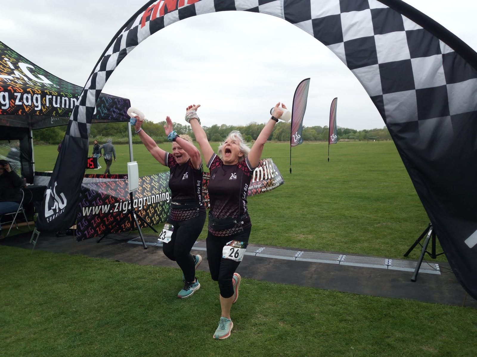 Two women are crossing a race finish line with their arms raised in victory. They are dressed in athletic gear, wearing black tops and leggings, and smiling excitedly. The finish line features a black-and-white checkered banner, and there are race-related tents and flags in the background.