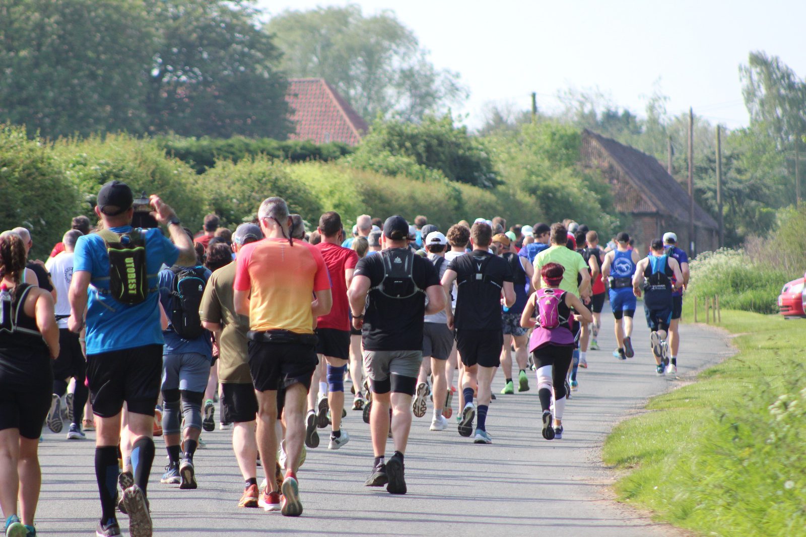 A large group of runners, wearing athletic wear and backpacks, participates in an outdoor race on a paved road surrounded by greenery. The runners are moving away from the camera, and some are throwing their arms in the air. The sky is clear and sunny.