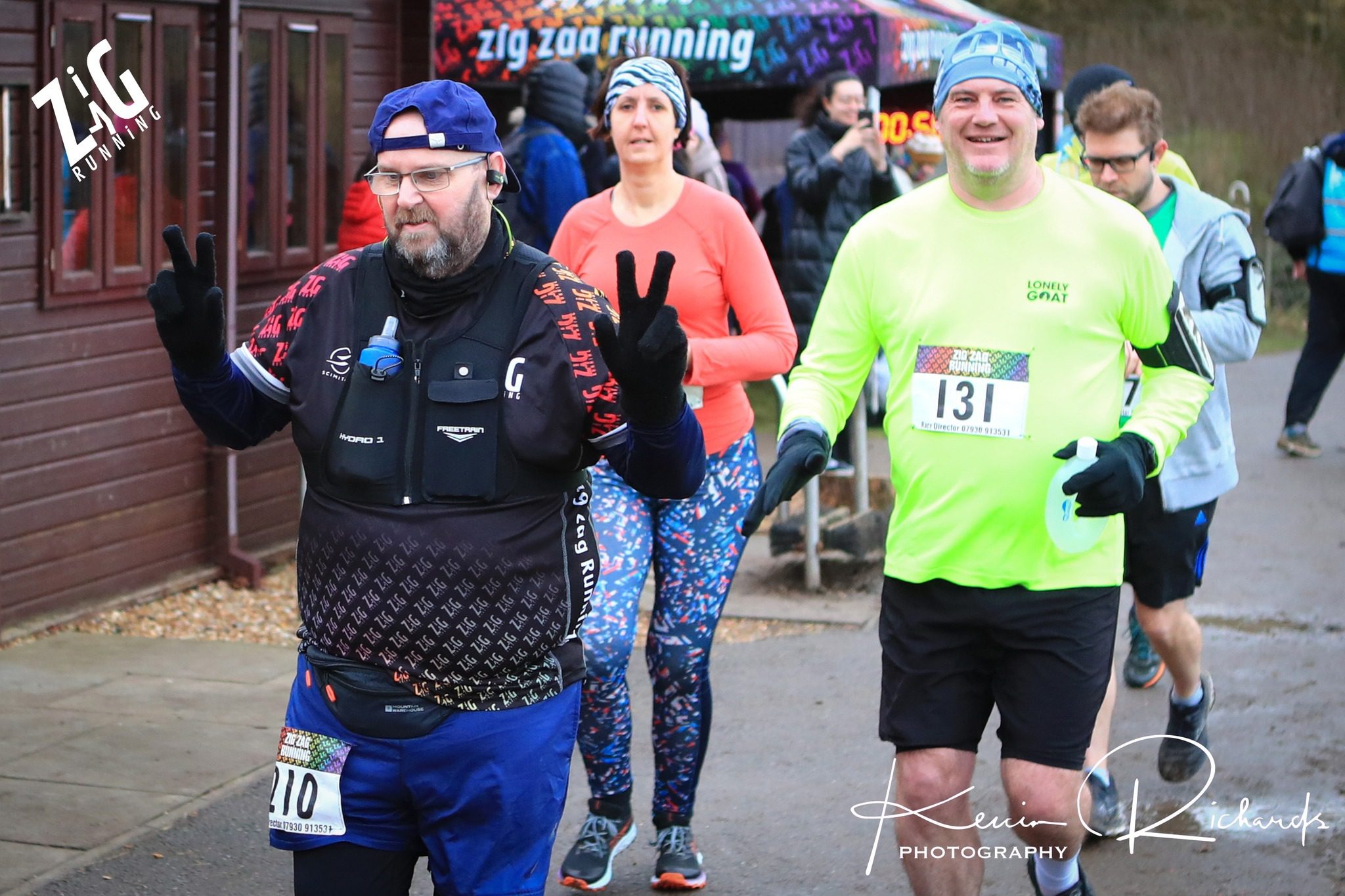 A group of runners participates in an outdoor race. One man, wearing a black vest and blue headband, gives a peace sign with both hands. Another man in a neon green shirt smiles. A woman in an orange top is behind them. They each sport race bibs and appear happy.