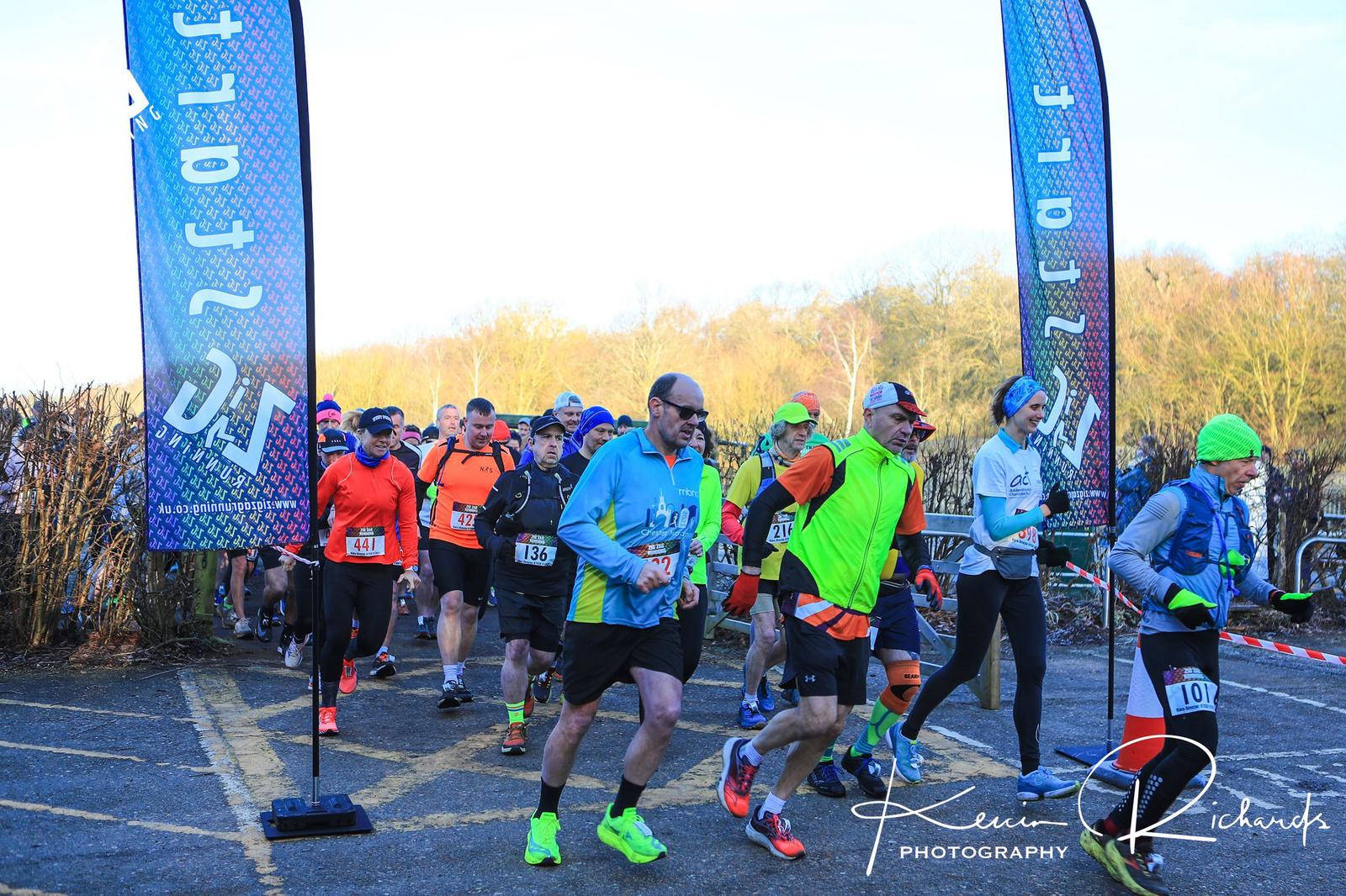 A group of runners in colorful attire, including neon and winter gear, participate in an outdoor race on a clear day. Two banners frame the starting area, and leafless trees are visible in the background. A sign reads "azit 202.
