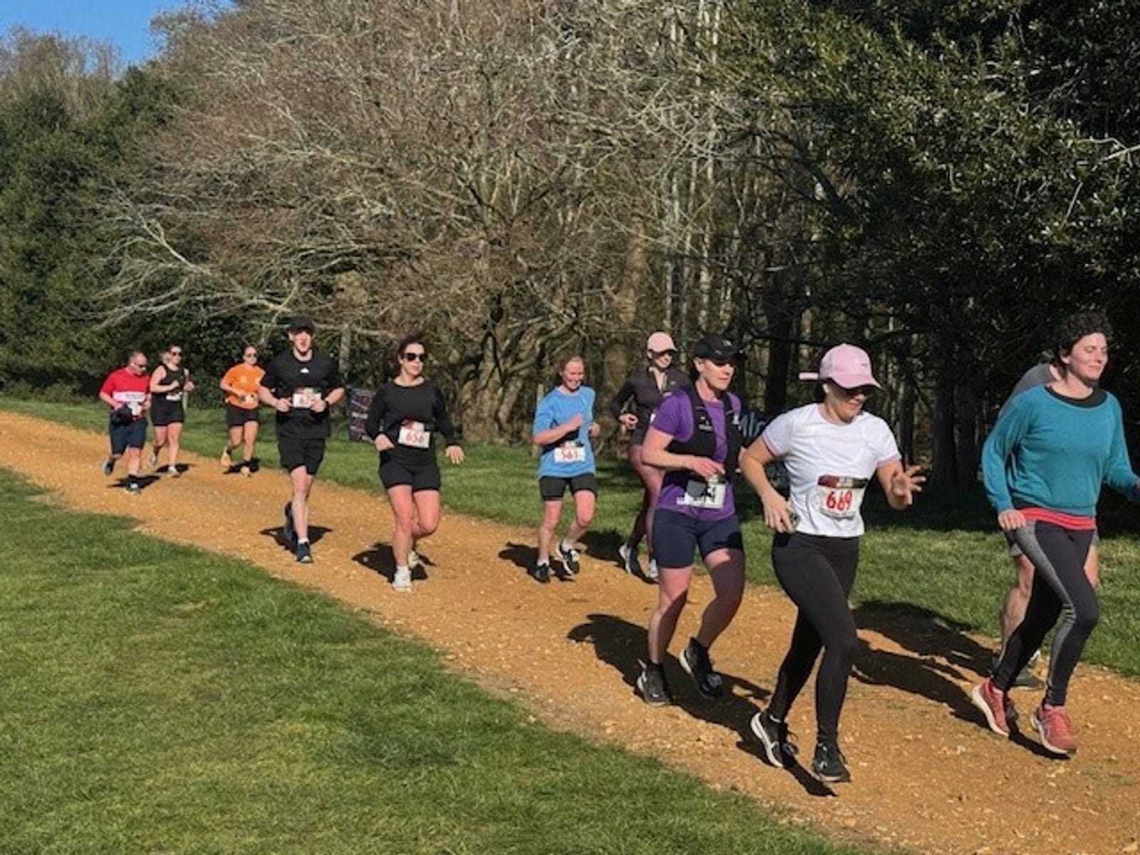 A group of runners, wearing numbered bibs and athletic gear, jog on a sunny dirt trail bordered by grass and trees. The runners vary in age and gender, and some wear hats or sunglasses.