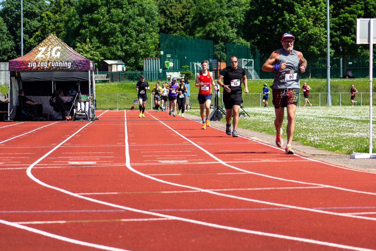 A group of runners in athletic gear competes on a red track under a sunny sky. A tent with the logo "Zig Zag Running" is visible on the left. Participants are at different stages, with one leading in the foreground. Green trees and grass surround the track.