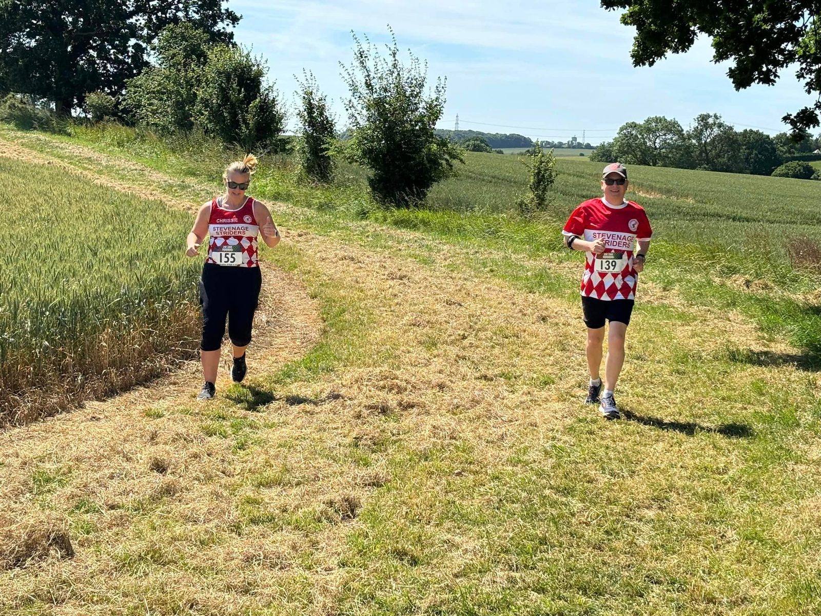 Two runners wearing red and white shirts with race numbers are jogging on a grassy path through a rural area. They are surrounded by fields and trees under a clear blue sky.