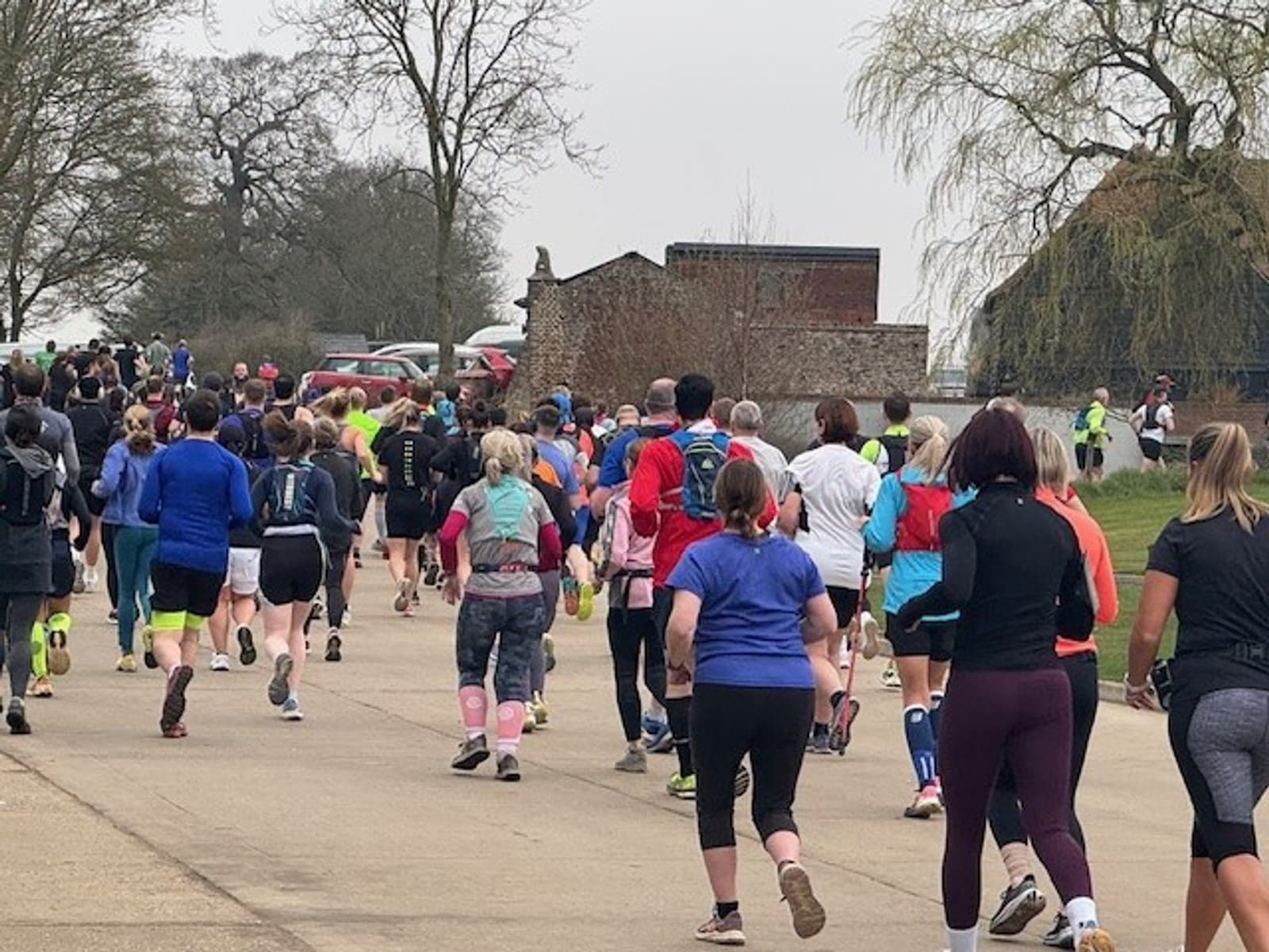 A group of people running on a paved path during a race or event. They are wearing athletic clothing and gear. Trees and a large building are visible in the background under a cloudy sky.