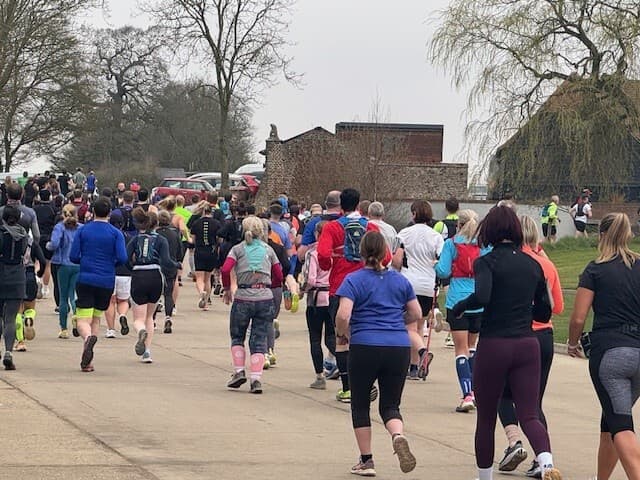 A group of people running on a paved path during a race or event. They are wearing athletic clothing and gear. Trees and a large building are visible in the background under a cloudy sky.