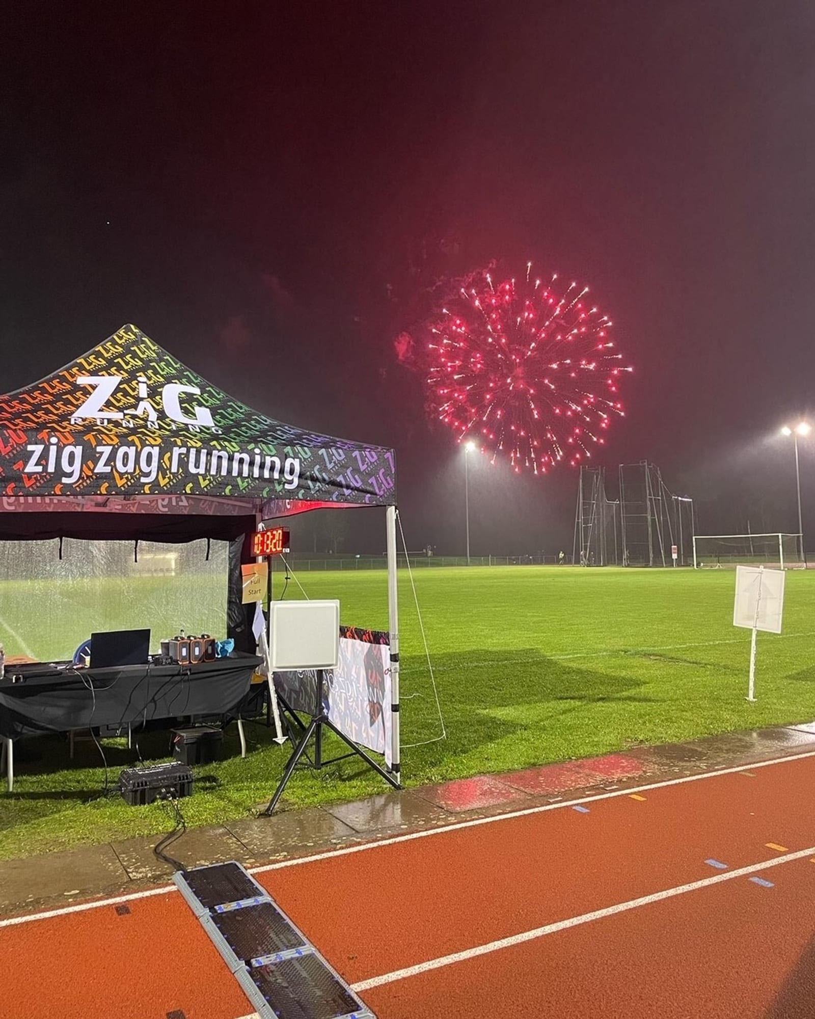 A running event tent labeled "zig zag running" is set up beside a wet athletics track at night, with bright red fireworks lighting up the sky in the background over a grassy field.