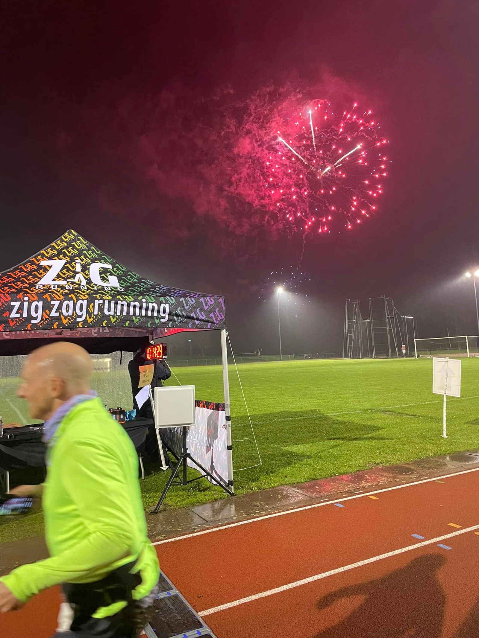 A runner in a green jacket is captured in motion on a wet track. A colorful tent marked "zig zag running" is visible, with a clock displaying race time in the background. Fireworks light up the night sky in shades of red and white over the sports field.