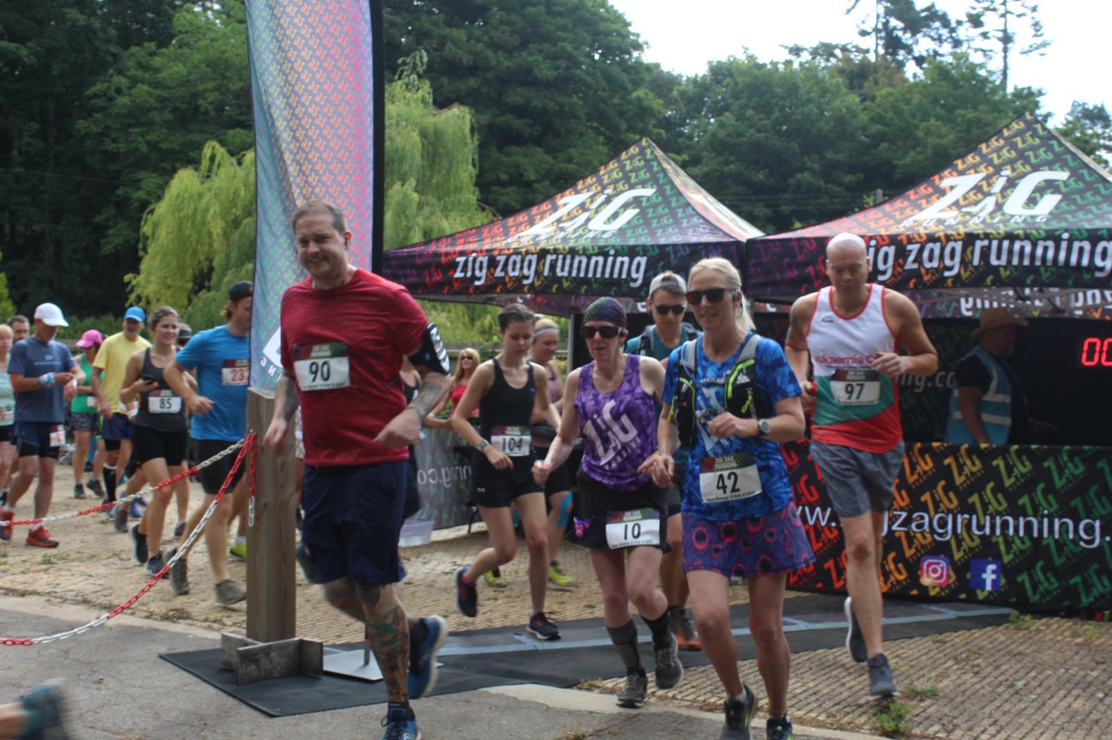A group of runners participating in a race, starting from a point marked by zigzag branded tents. The racers are diverse in attire and number tags, with a backdrop of greenery and trees.