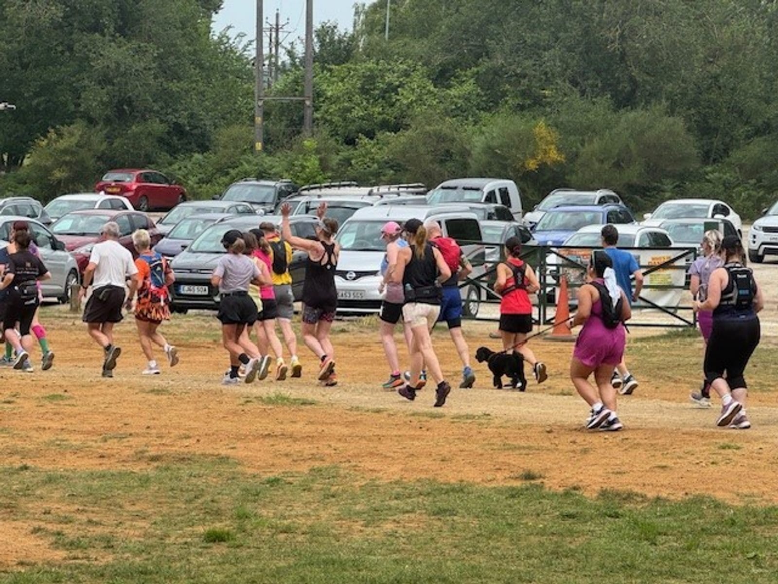 A group of people jog together on a dirt path in a park, with several parked cars and trees in the background. Most are wearing athletic clothing and backpacks, and one person is running with a small black dog.