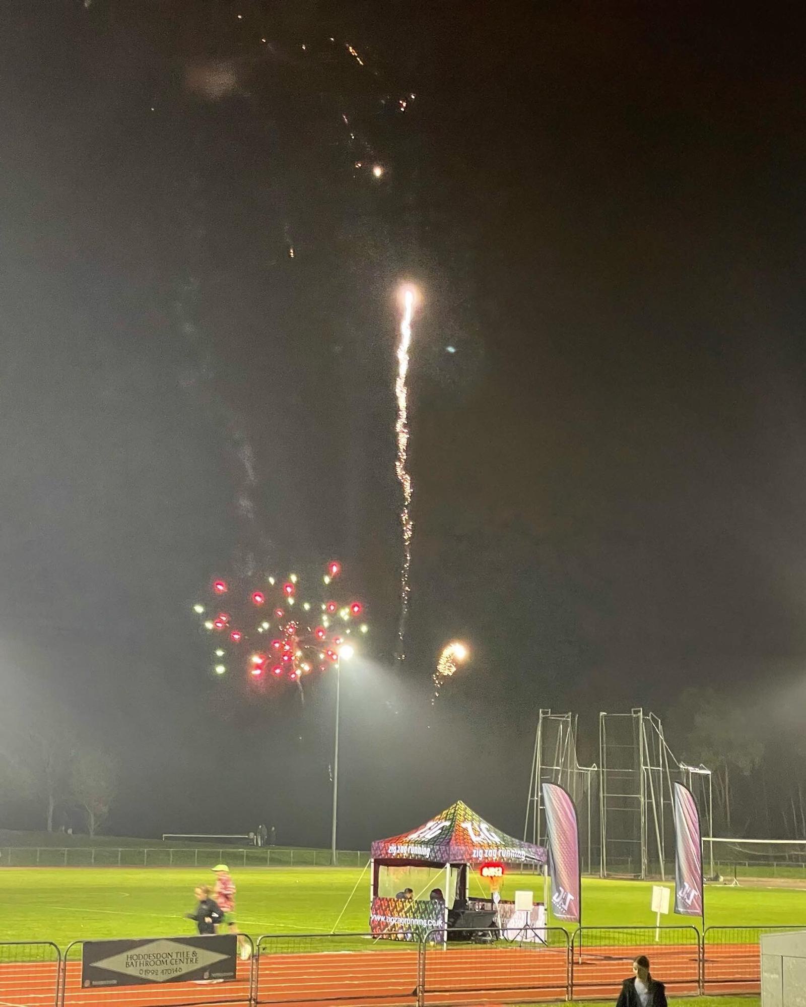 A nighttime scene at a sports field features a small tent with colorful fabric and banners on either side. Fireworks are exploding in the sky above, creating bright, colorful patterns. A few people are visible in the foreground, and the field is dimly lit.