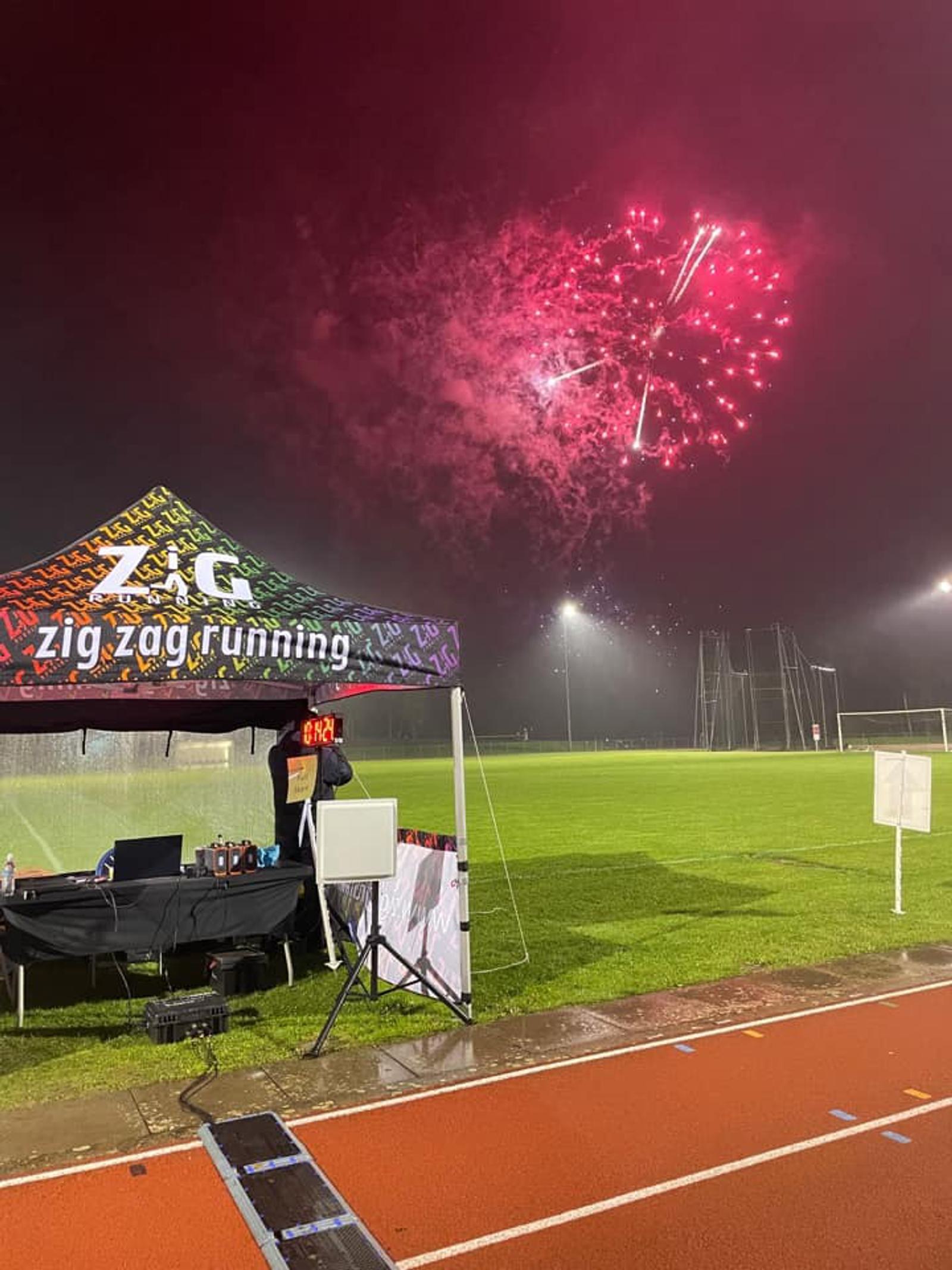 A nighttime scene featuring a sports field illuminated by bright stadium lights. Colorful fireworks burst in the sky. In the foreground, a "Zig Zag Running" tent stands with a person beside it and a red running track is visible beneath.