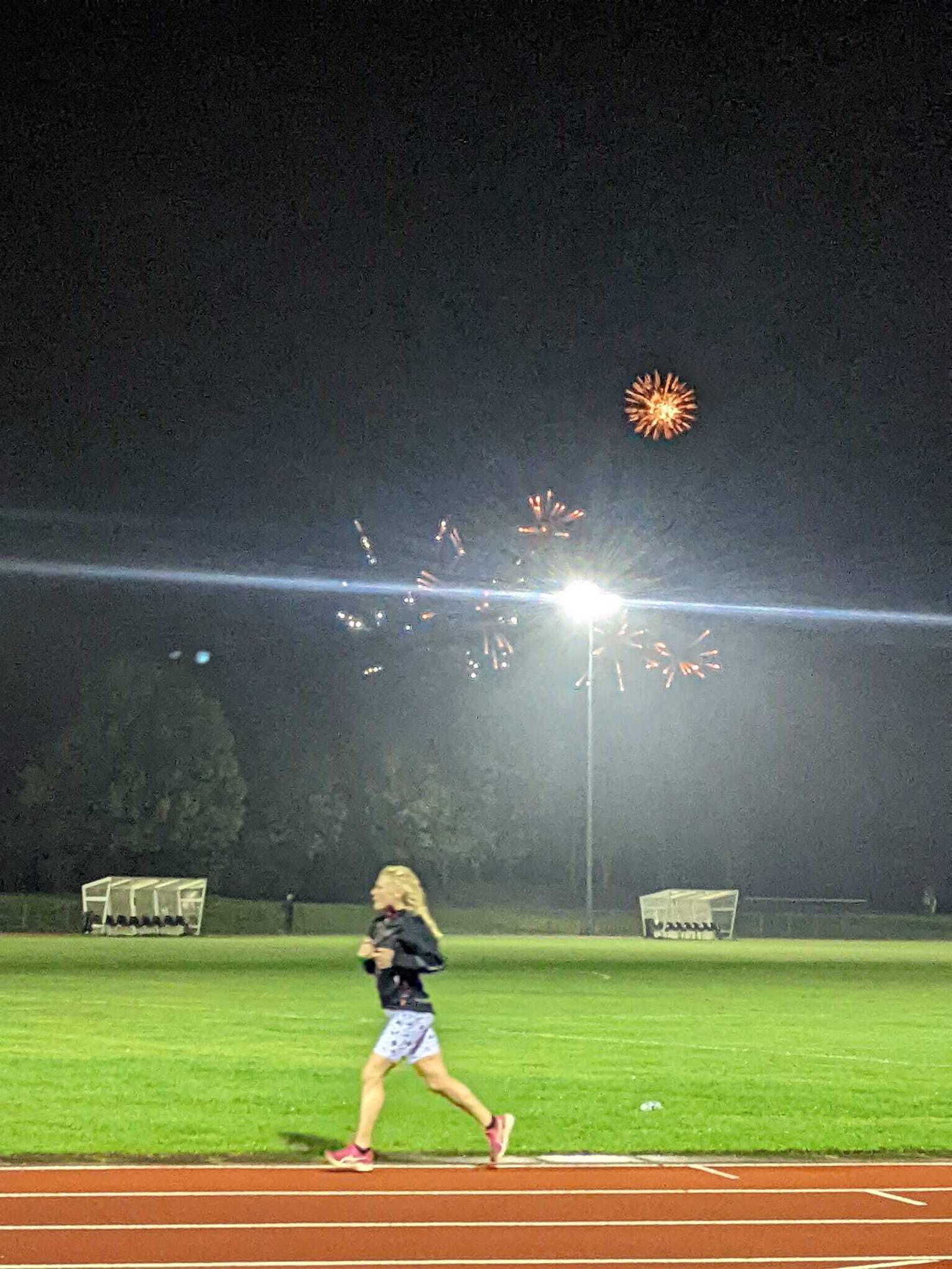 A person with blonde hair is walking on a sports track at night. There are bright fireworks bursting in the sky in the background, illuminating the scene. The grassy field beside the track has benches or shelters under a floodlight.