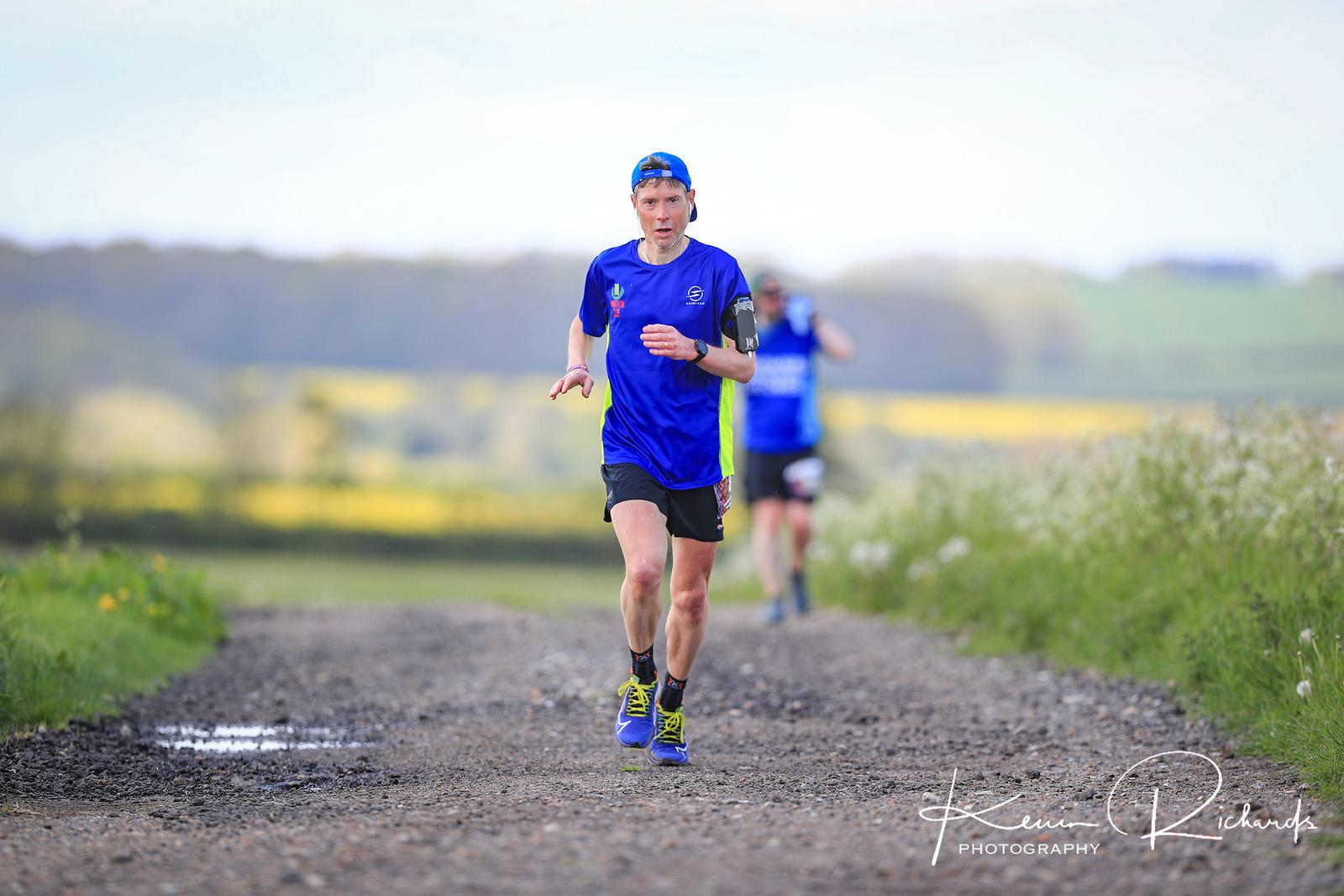 A runner in a blue shirt and cap races along a gravel path in a rural area, with a blurred background of fields and greenery. Another runner is visible in the distance behind him. The photo is credited to Kevin Richards Photography.