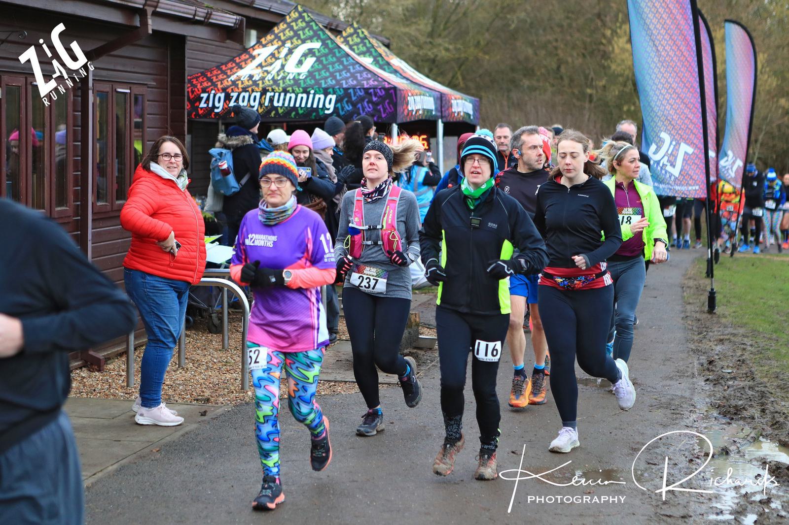 A group of runners in colorful attire participate in a race, passing a wooden building with a "zig zag running" banner. Some spectators watch from the sidelines. The background features trees and two flags.