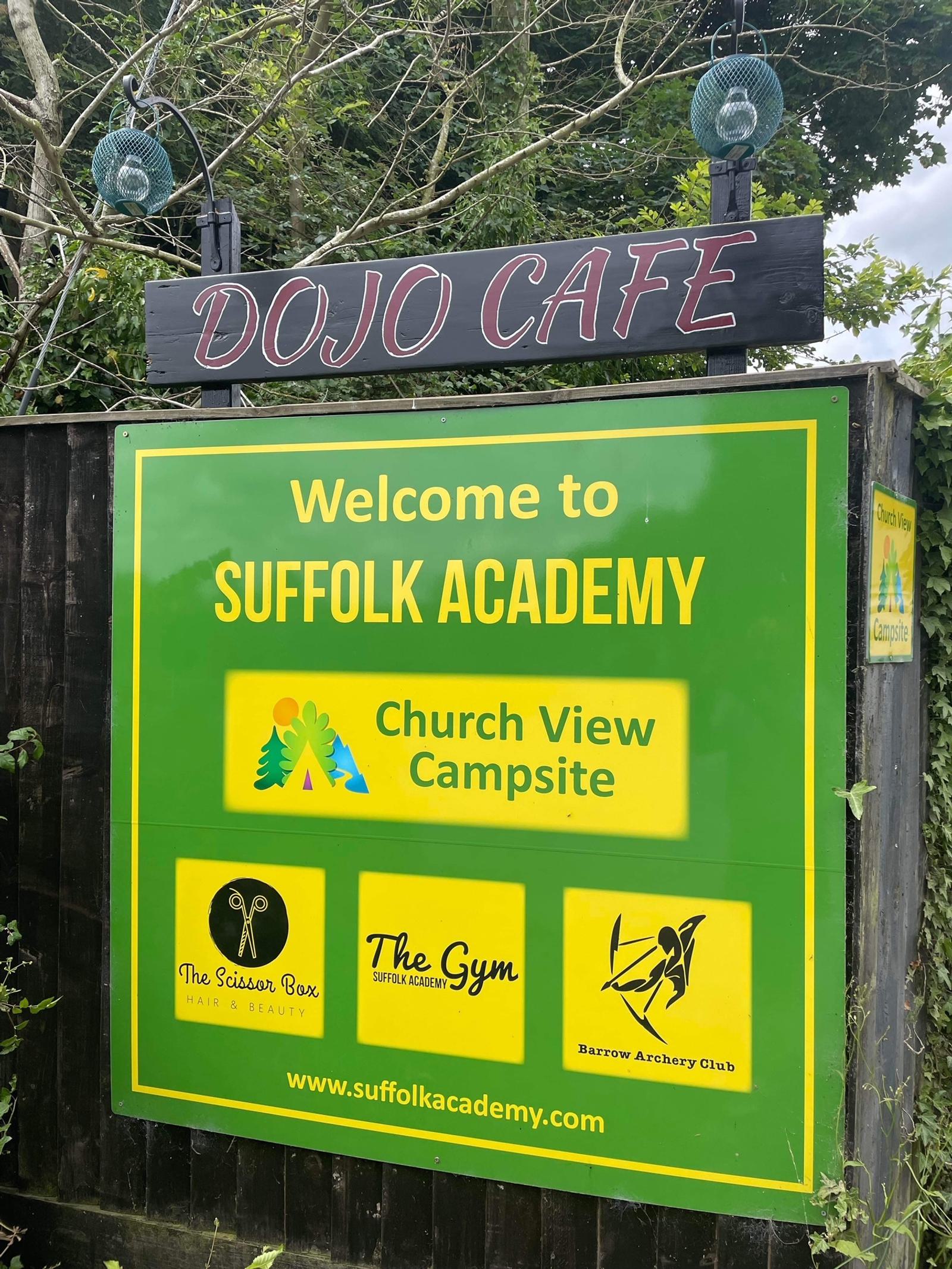 A sign for Suffolk Academy, located at Church View Campsite, with a green background. It features logos for The Scissor Box Hair & Beauty, The Gym Suffolk Academy, and Barrow Archery Club. Above the sign is a smaller sign for DOJO CAFE. Trees are visible in the background.